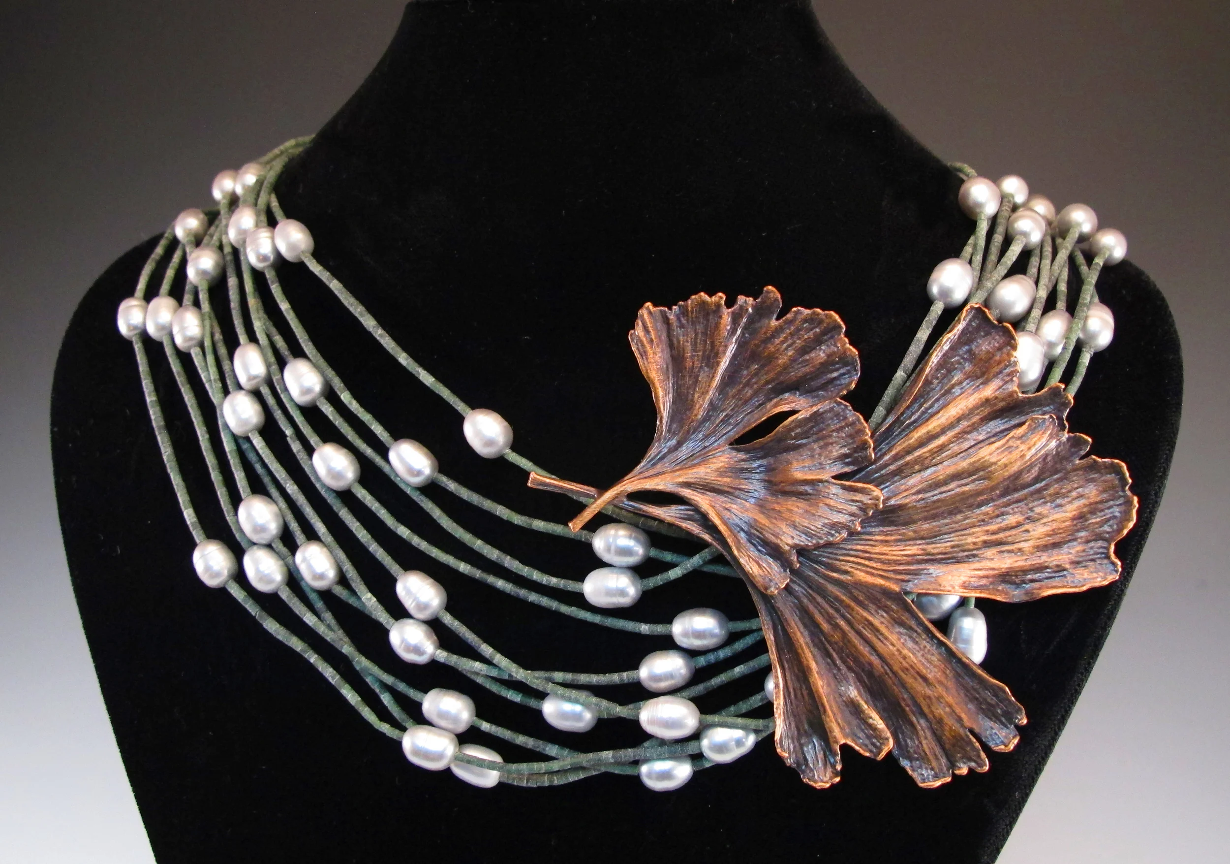 A layered pearl necklace with strands of gray cords, large white pearls, and a copper-colored leaf pendant, displayed on a black velvet bust