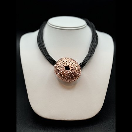 A necklace with a large, round, textured, rose gold pendant on a black multi-strand cord, displayed on a white jewelry bust.