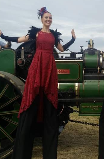 A woman standing in front of a vintage green steam locomotive, smiling, wearing a red and black dress with feathered accents.