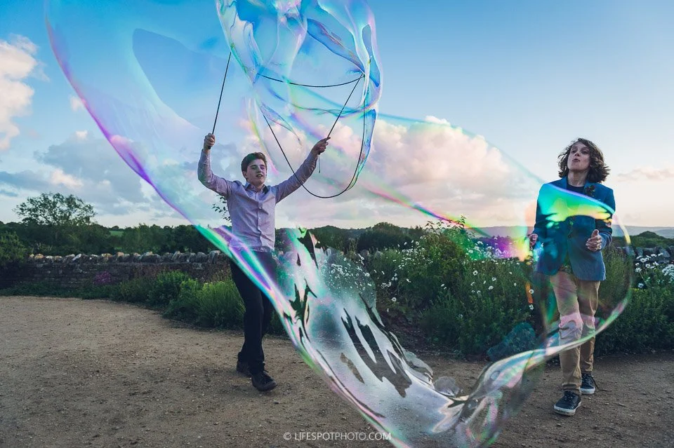 Two kids playing with a large soap bubble outdoors on a dirt path with greenery and a cloudy sky in the background.