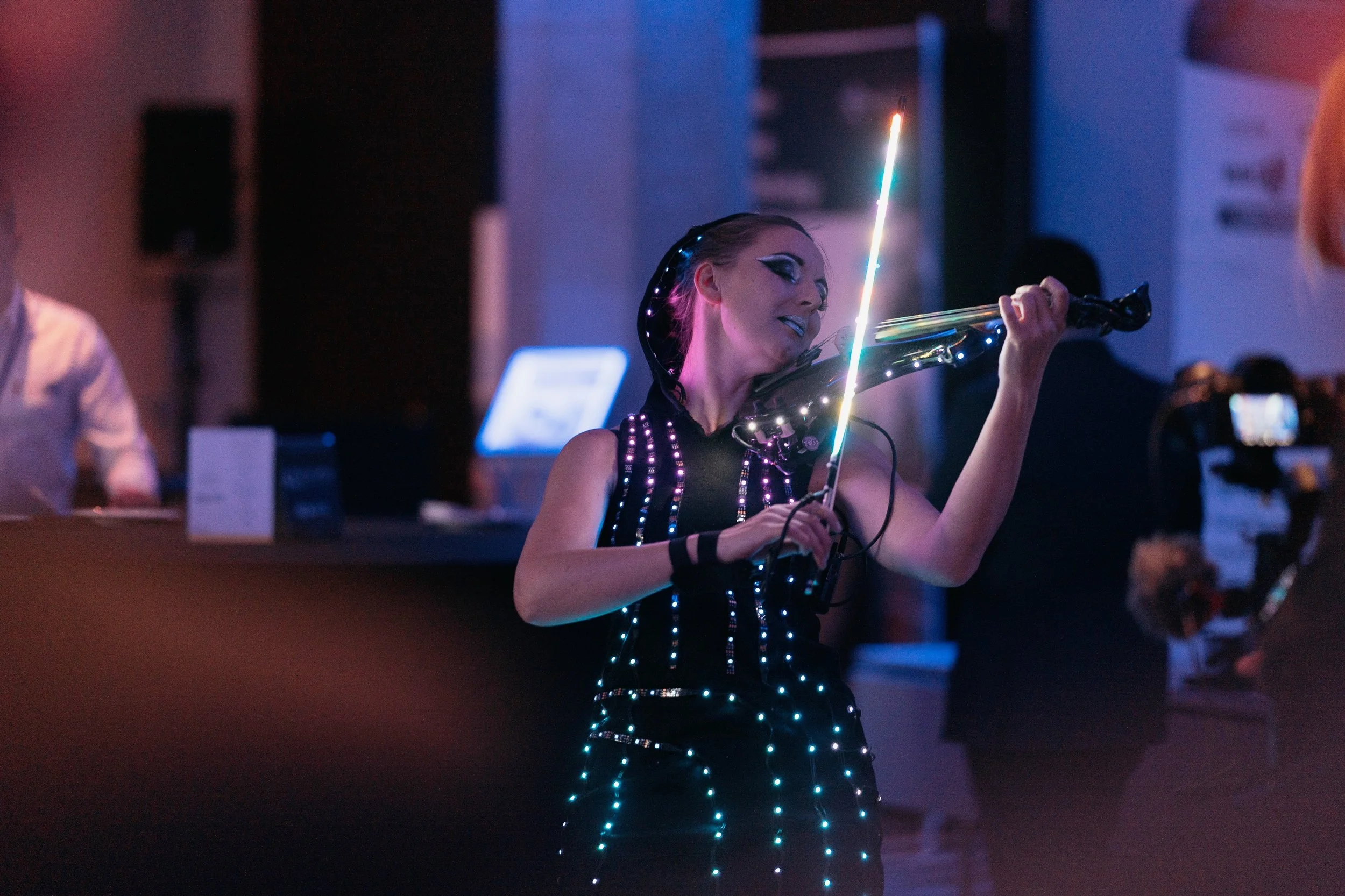 Woman playing the violin at a dimly lit event, wearing a dress decorated with LED lights, with colorful light effects and a dark background.