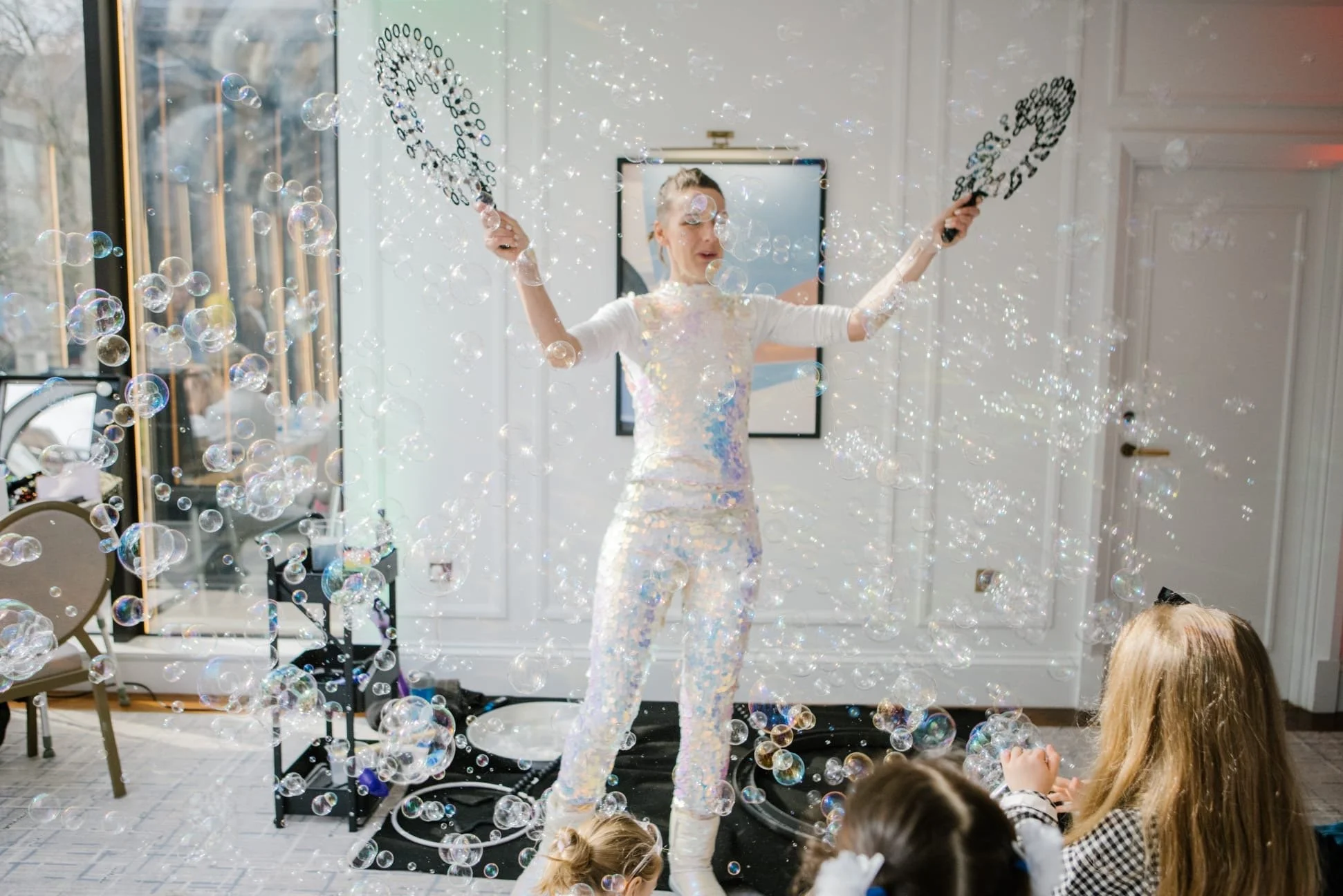A woman dressed in a shiny, iridescent outfit performs a bubble show for children in a brightly lit room while some children watch and play nearby.