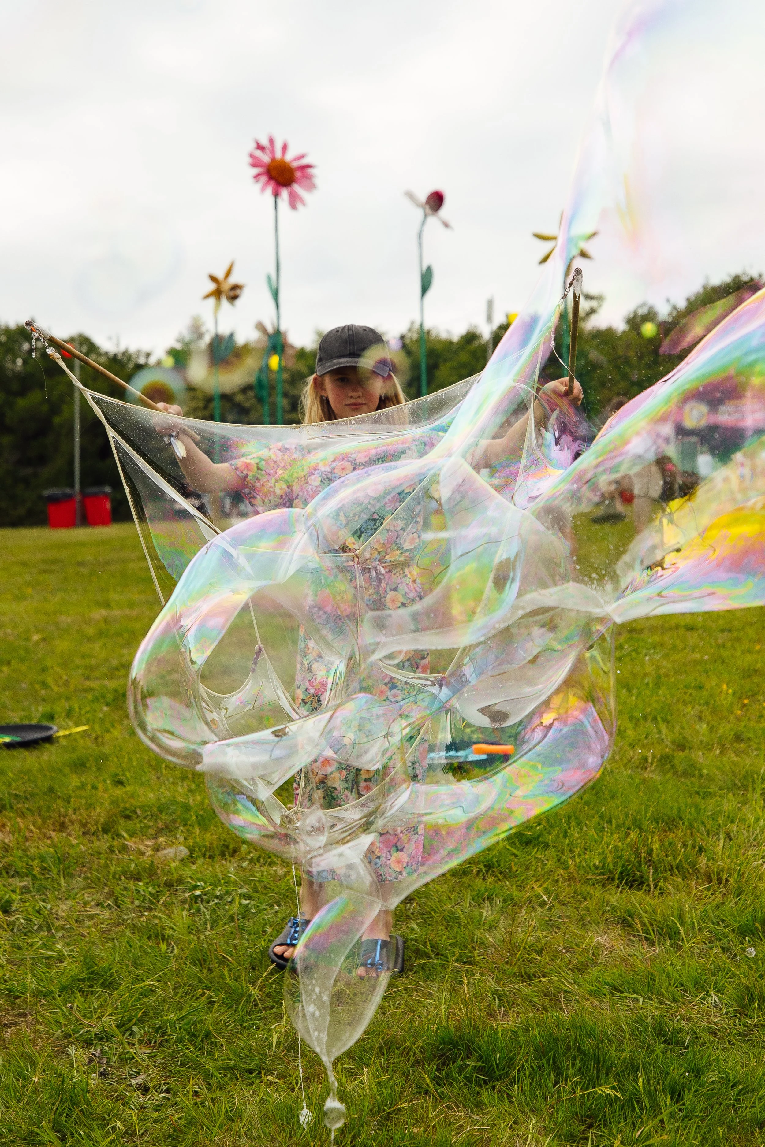 A girl in a floral dress and black cap playing with large, colorful soap bubbles outdoors on grass, with tall flower sculptures in the background under an overcast sky.