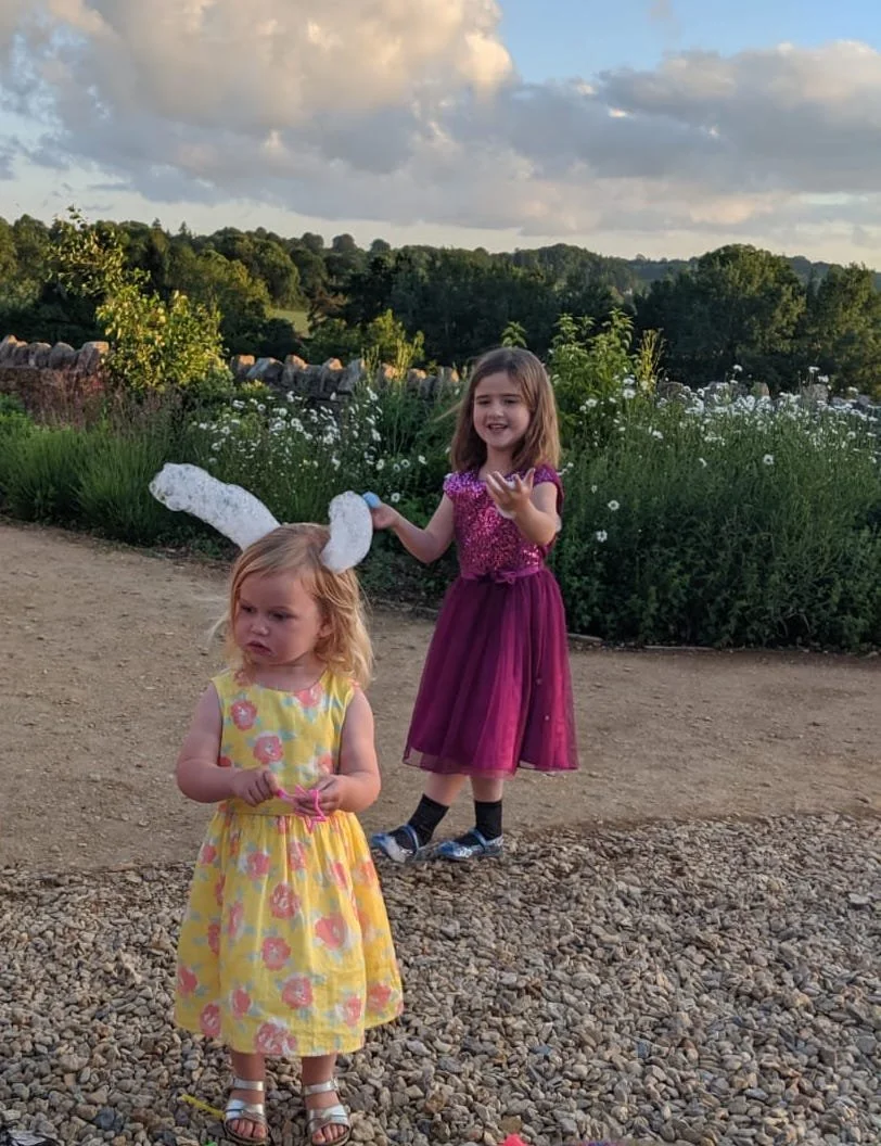 Two young girls are outdoors on a dirt path with a garden and trees in the background under a cloudy sky. One girl, with blonde hair, is wearing a sleeveless yellow dress with pink flowers, white sandals, and bunny ear headband, and looks serious. Th