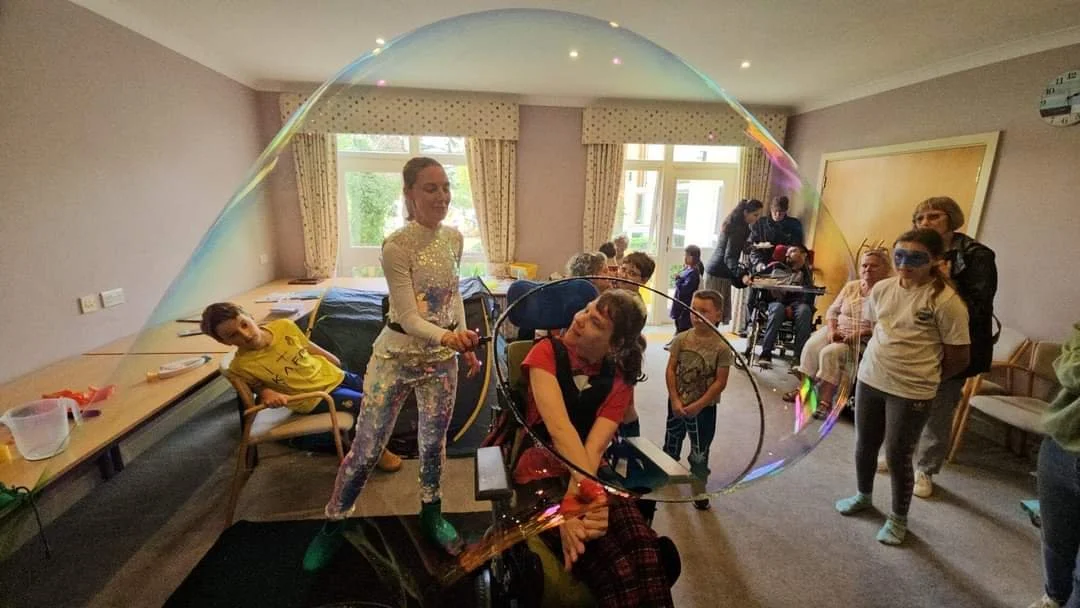 A woman making a large soap bubble with children and adults in a room with beige walls and windows.