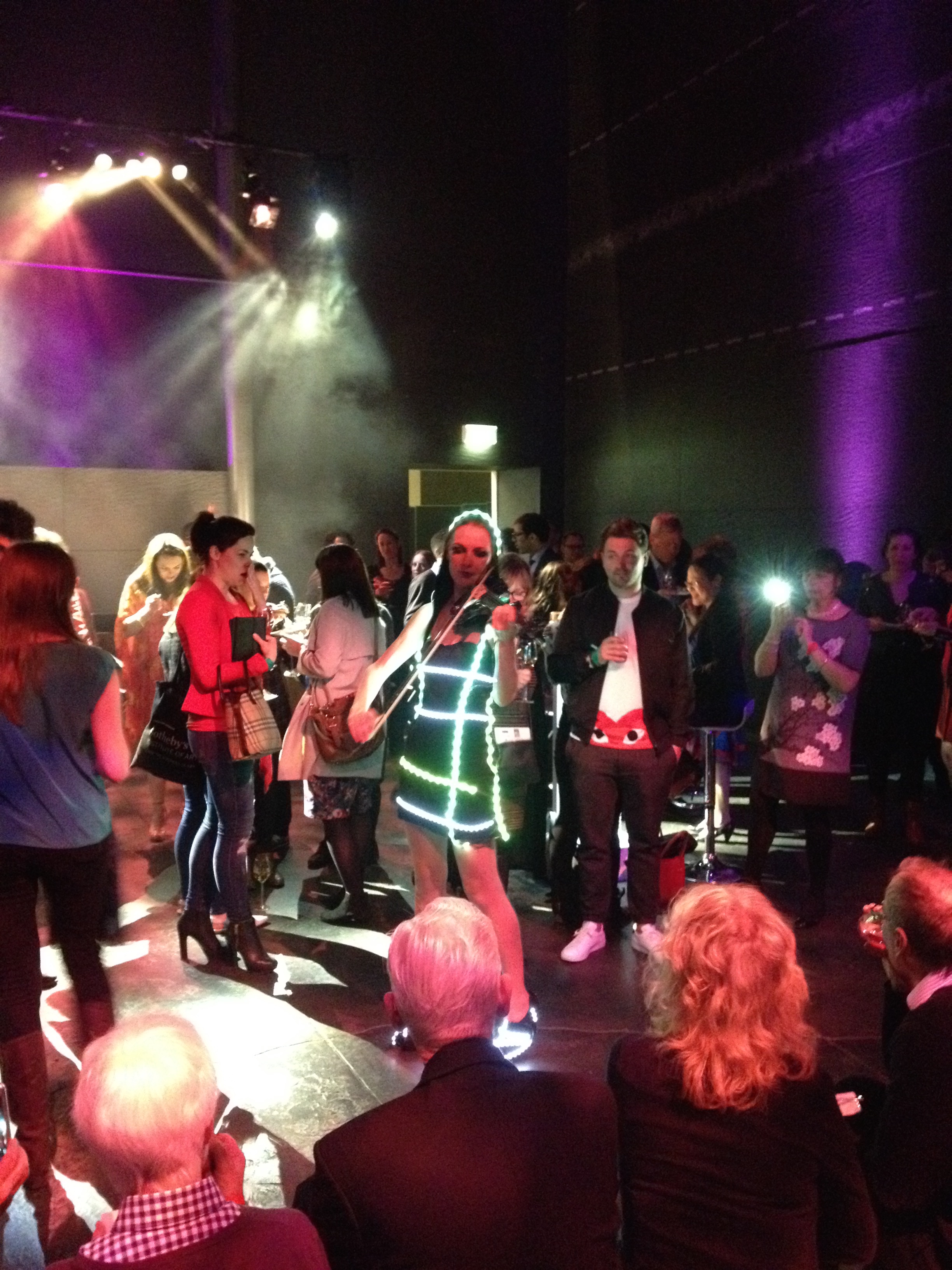 People gathered at a lively indoor event with colorful stage lighting, some individuals holding drinks, and others using their phones among dark walls and ceiling.