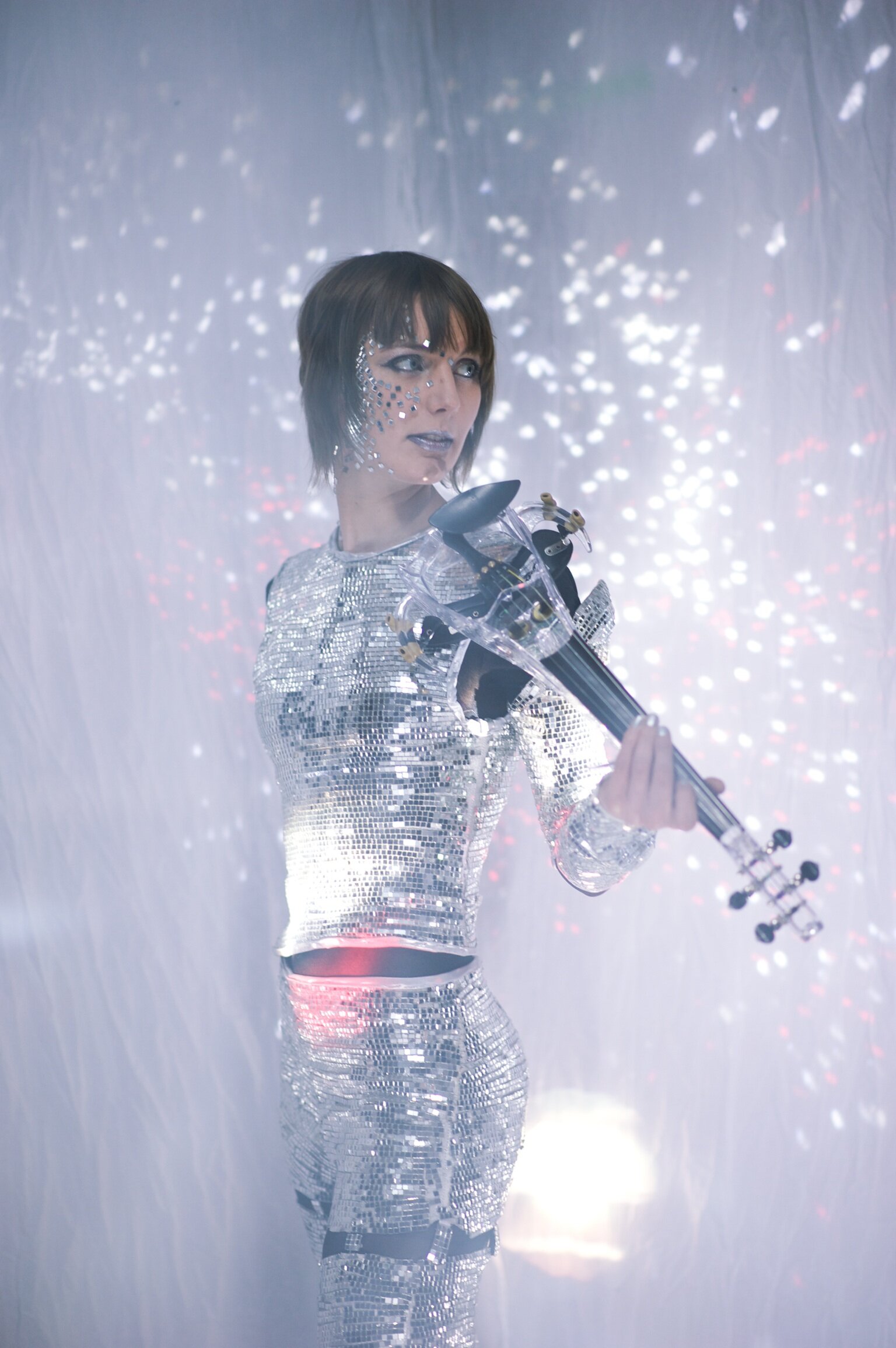 Person dressed in a shiny, reflective mirror outfit playing a transparent violin with a background of sparkling light effects resembling stars or fireworks.