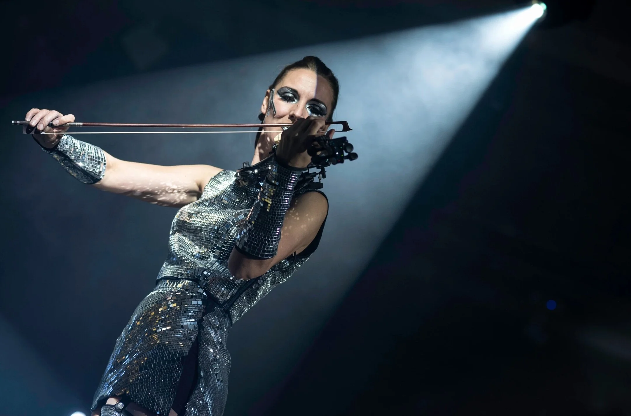 A woman performing on stage wearing a shiny, mirror-like outfit and holding a bow and arrow, with stage lighting creating a dramatic effect.