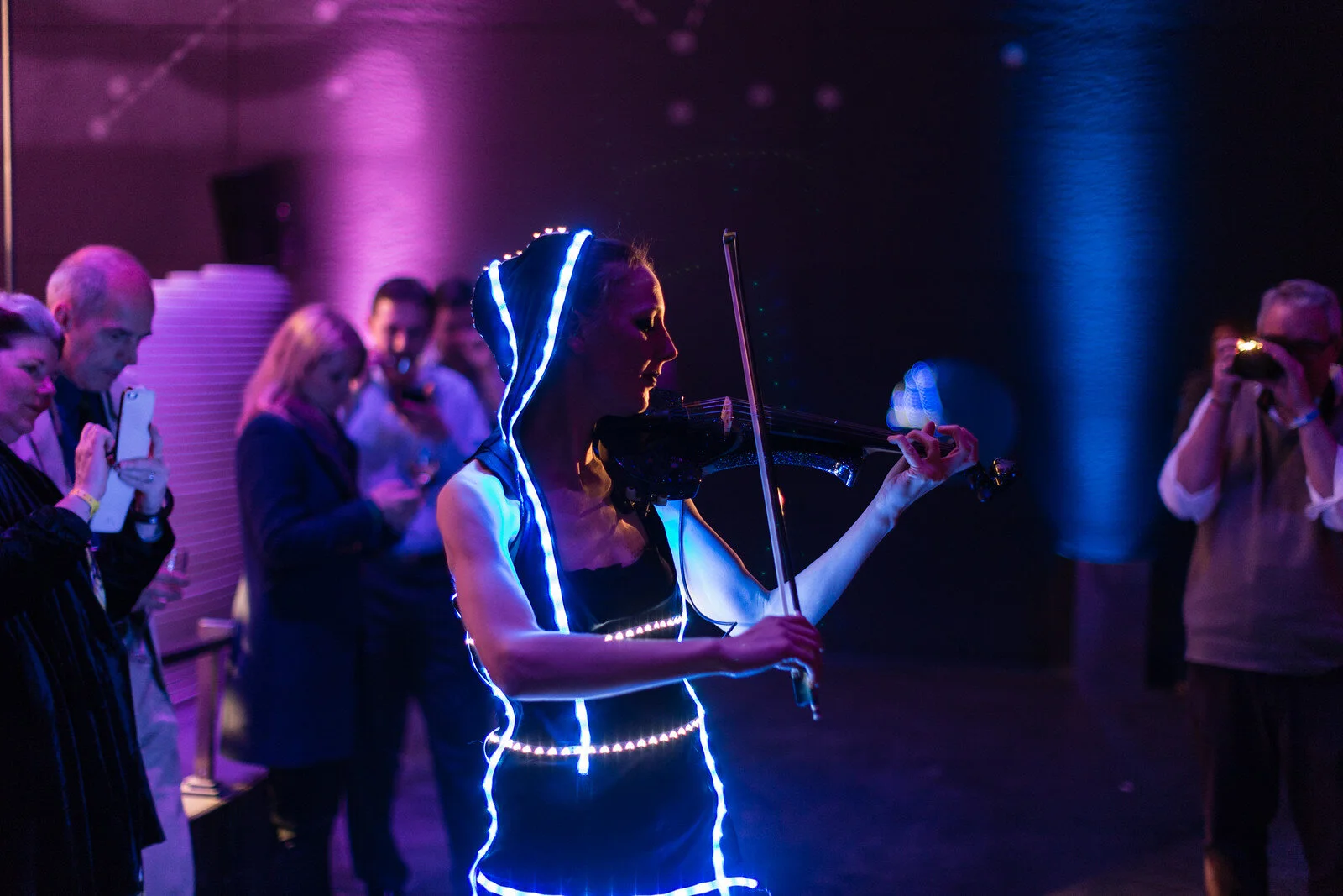 A woman dressed in black with glowing blue and white light strips is playing a violin at a dark, futuristic event. Several people are watching and taking pictures with phones.