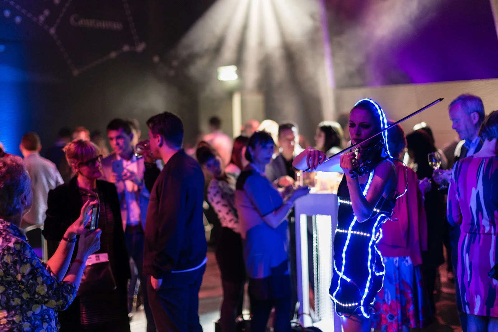 A woman dressed in a colorful outfit decorated with LED lights playing the violin at a crowded indoor event or party.