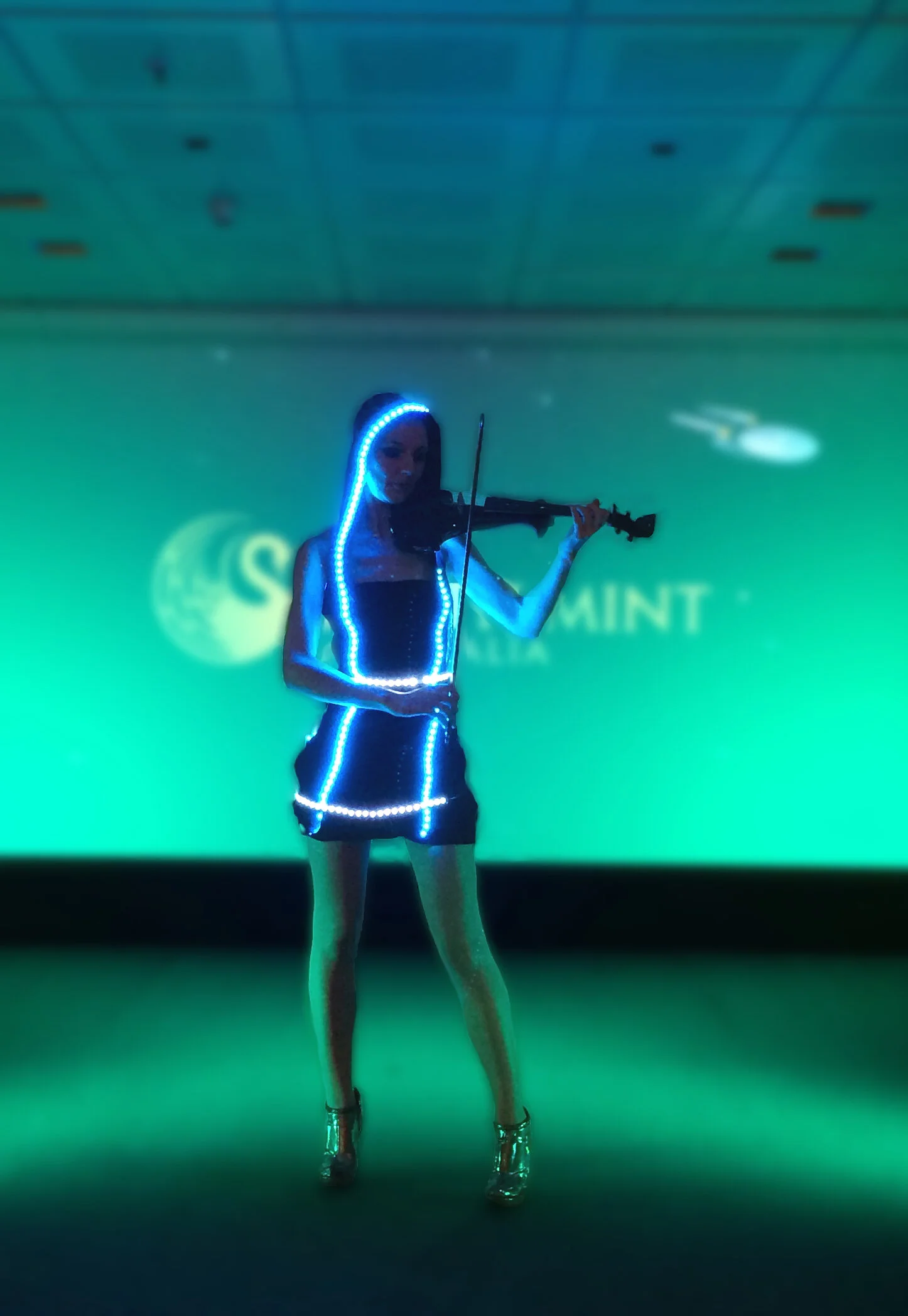 A woman playing a violin on a stage, wearing a black dress with LED lights along the edges, against a green background with blurred text.