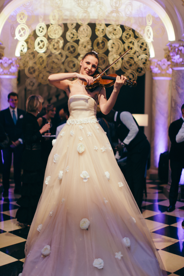 A woman in a wedding gown playing a violin at a wedding reception ballroom decorated with circular light fixtures and floral decor.