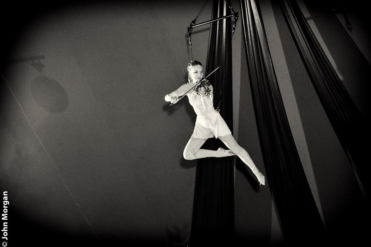 A female aerialist performing on a trapeze in a theater, with a focused expression and black curtains in the background.