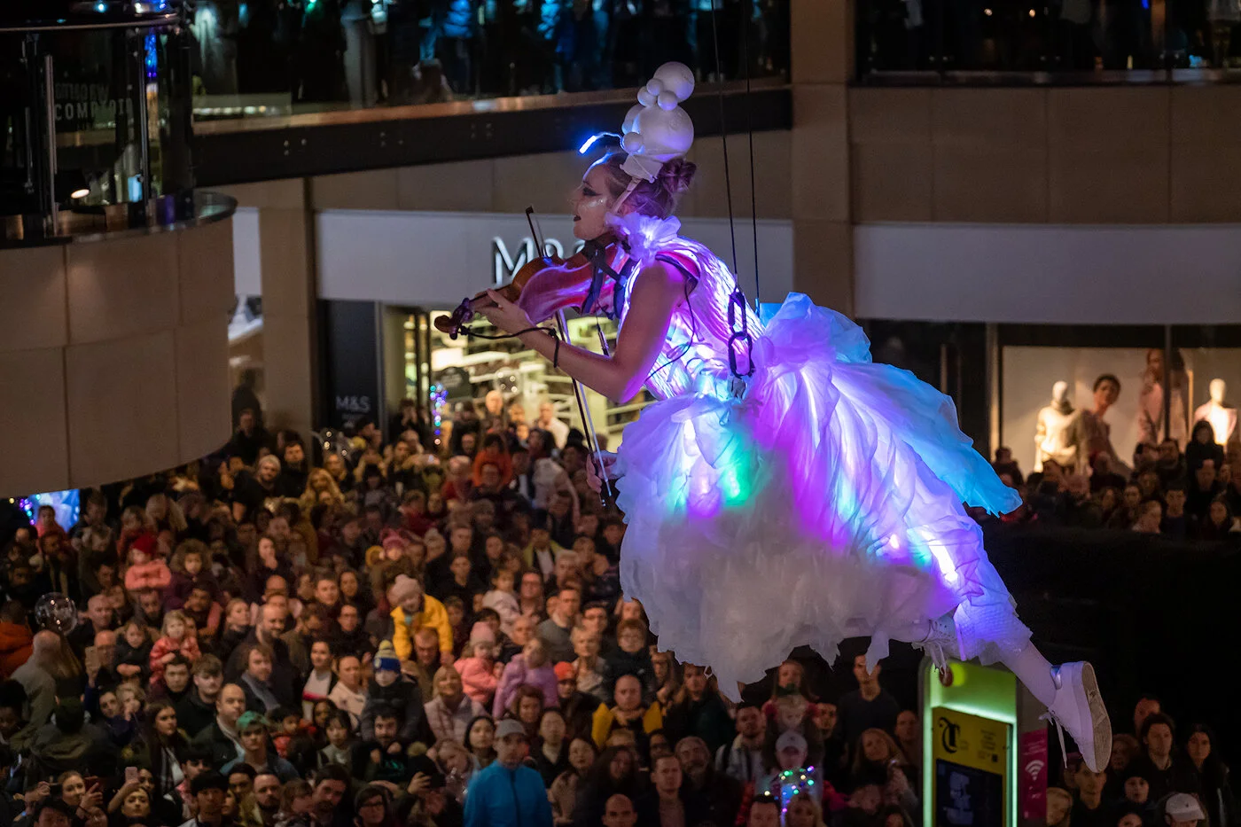 A performer dressed in a glowing, illuminated costume and headpiece, playing the violin above a crowd at a public event or parade.