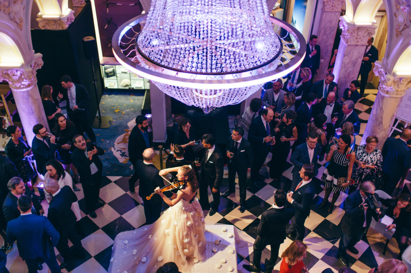 A lavish ballroom during an elegant event with a large crystal chandelier, guests in formal attire dancing, drinking, and socializing, with a woman in a flowing gown playing the violin.