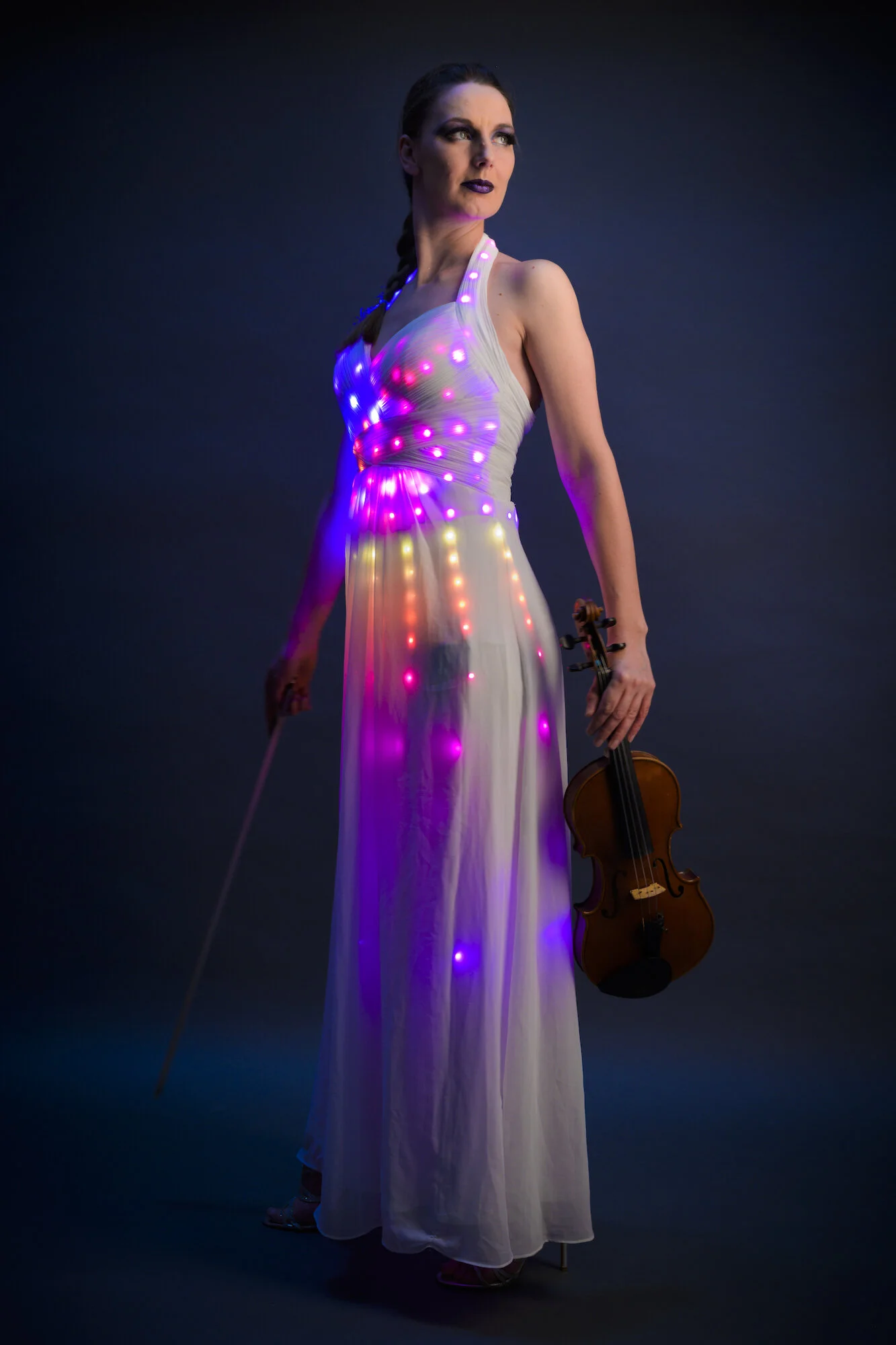 A woman in a flowy dress with LED lights, holding a violin, standing against a dark background.