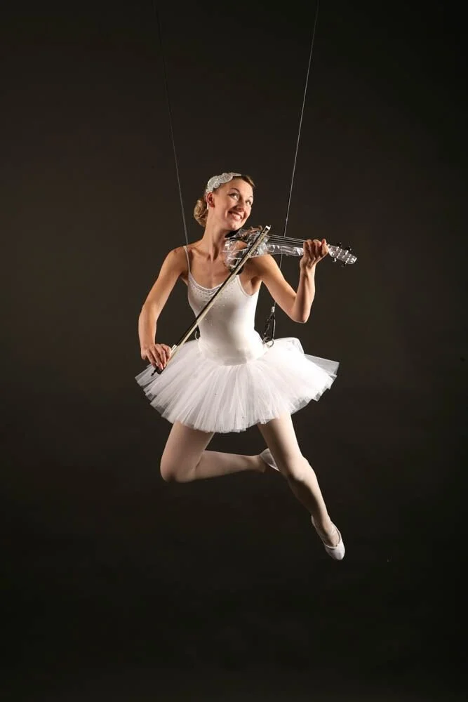 A woman in a ballet tutu and pointe shoes playing a transparent electric violin while suspended in mid-air, seen against a dark background.