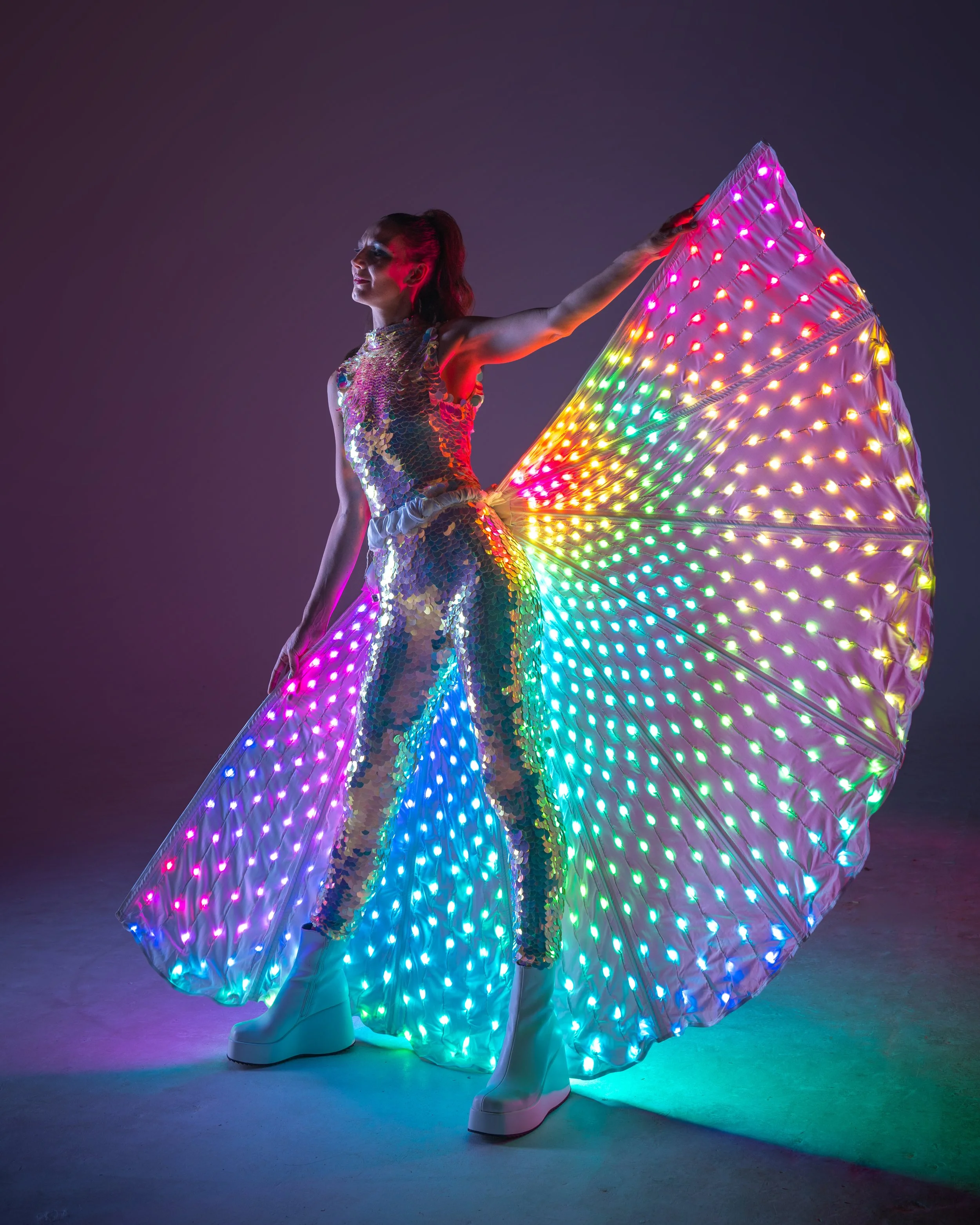 A person wearing a dress and boots, holding a large, illuminated, rainbow-colored umbrella with multicolored LED lights.