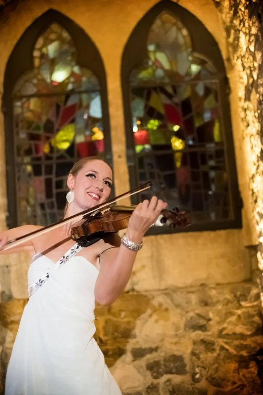 A woman in a white dress playing a violin in a warmly lit church with stained glass windows in the background.