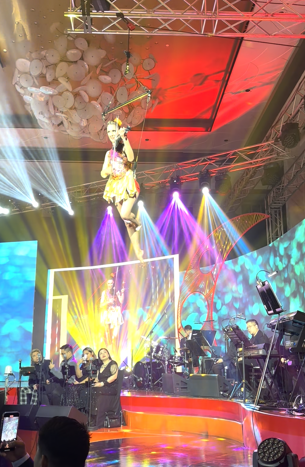 A stage with a performer hanging from a harness above the stage, surrounded by colorful lights and a band playing instruments, with an audience visible in front.