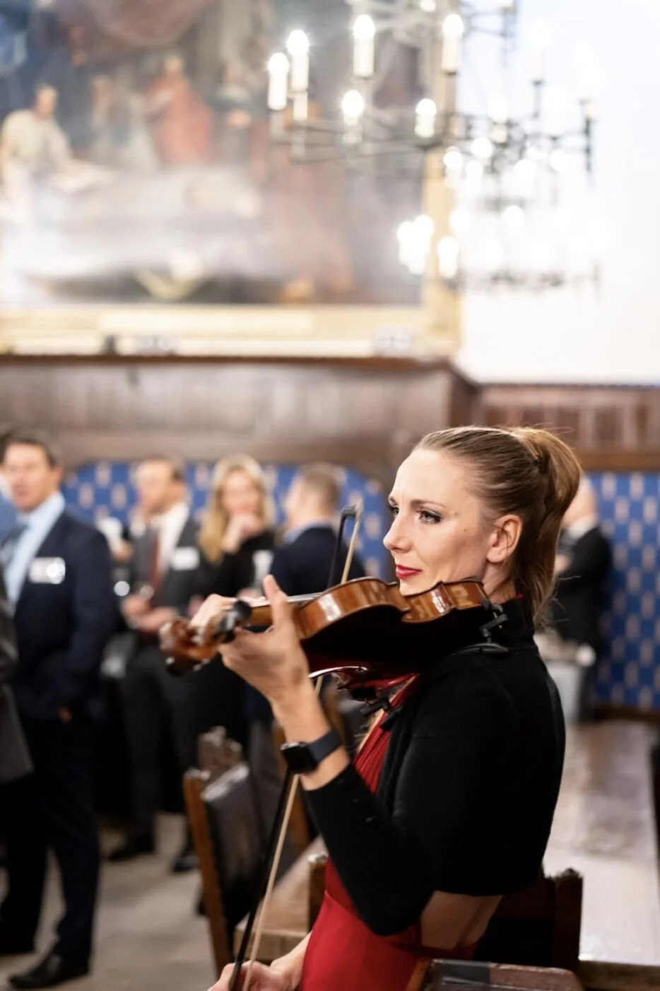 A woman playing the violin at a formal gathering in a room with wood-paneled walls and a large painting on the wall.