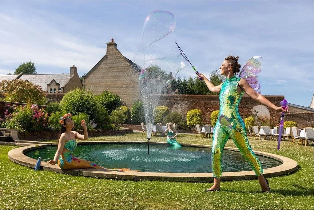 Two women dressed as mermaids and fairy wings playing with a bubble machine in a backyard garden with flowers, trees, and house in the background.