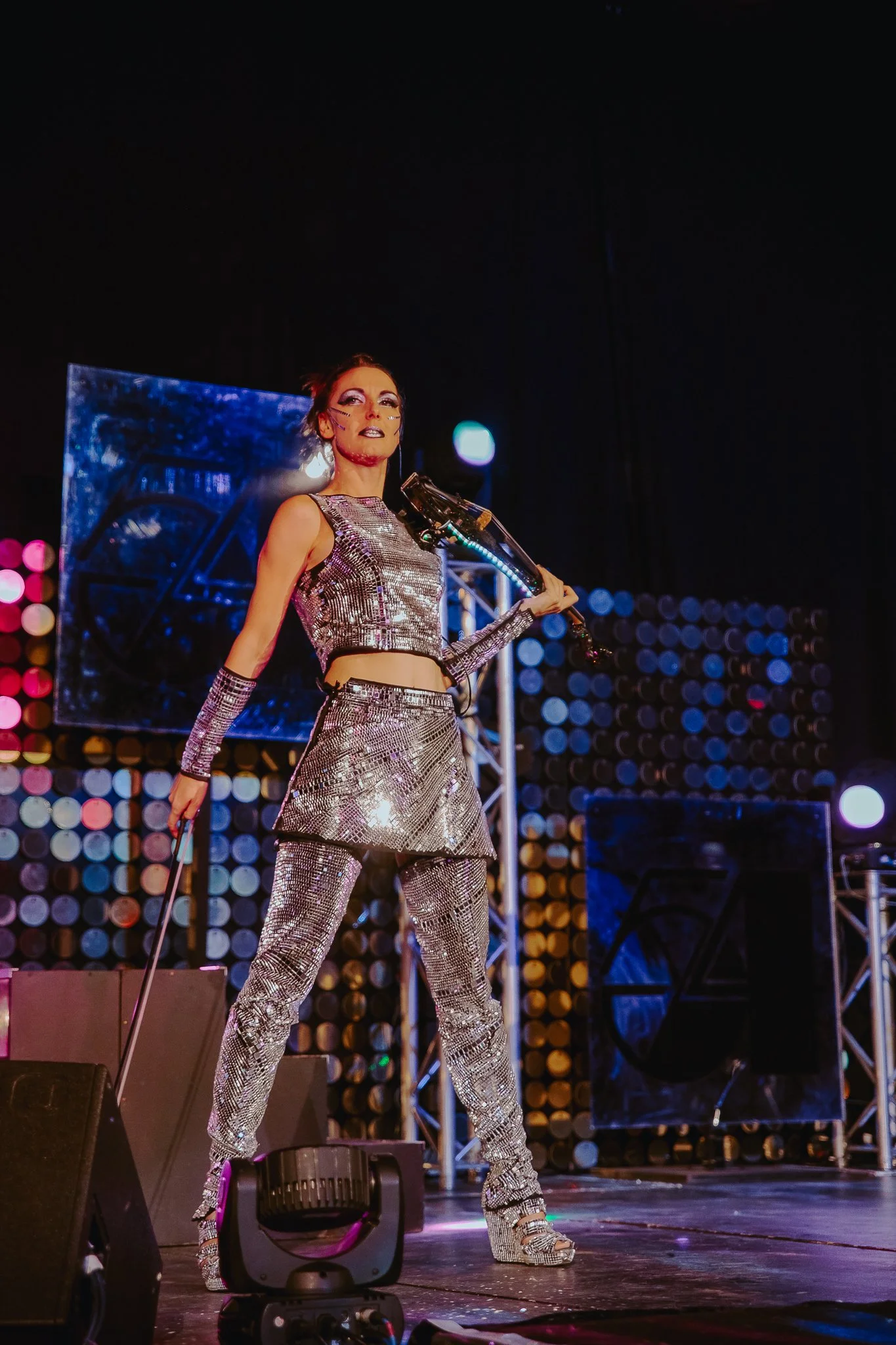 Performer on stage wearing a metallic silver crop top, matching pants, and high heels, holding a microphone, with stage lighting and background out of focus.