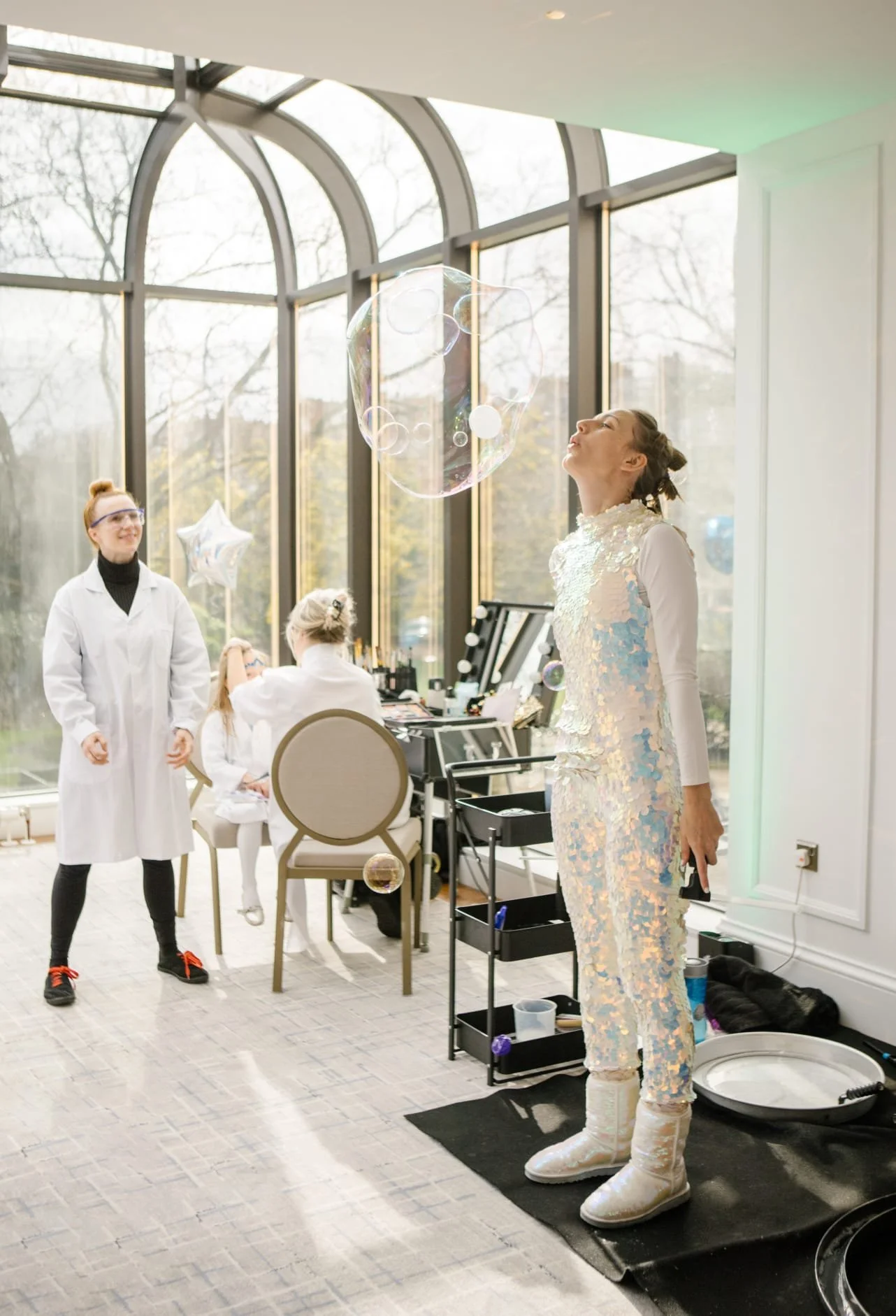 A woman with light skin in a shiny, iridescent outfit stands on a black mat, surrounded by a bubble, in a bright room with large windows. Two girls in white coats and glasses, one with a bun, and a third girl in a lab coat sit at a table in the background. The room has a makeup station and decorative balloons, indicating a festive or special event.