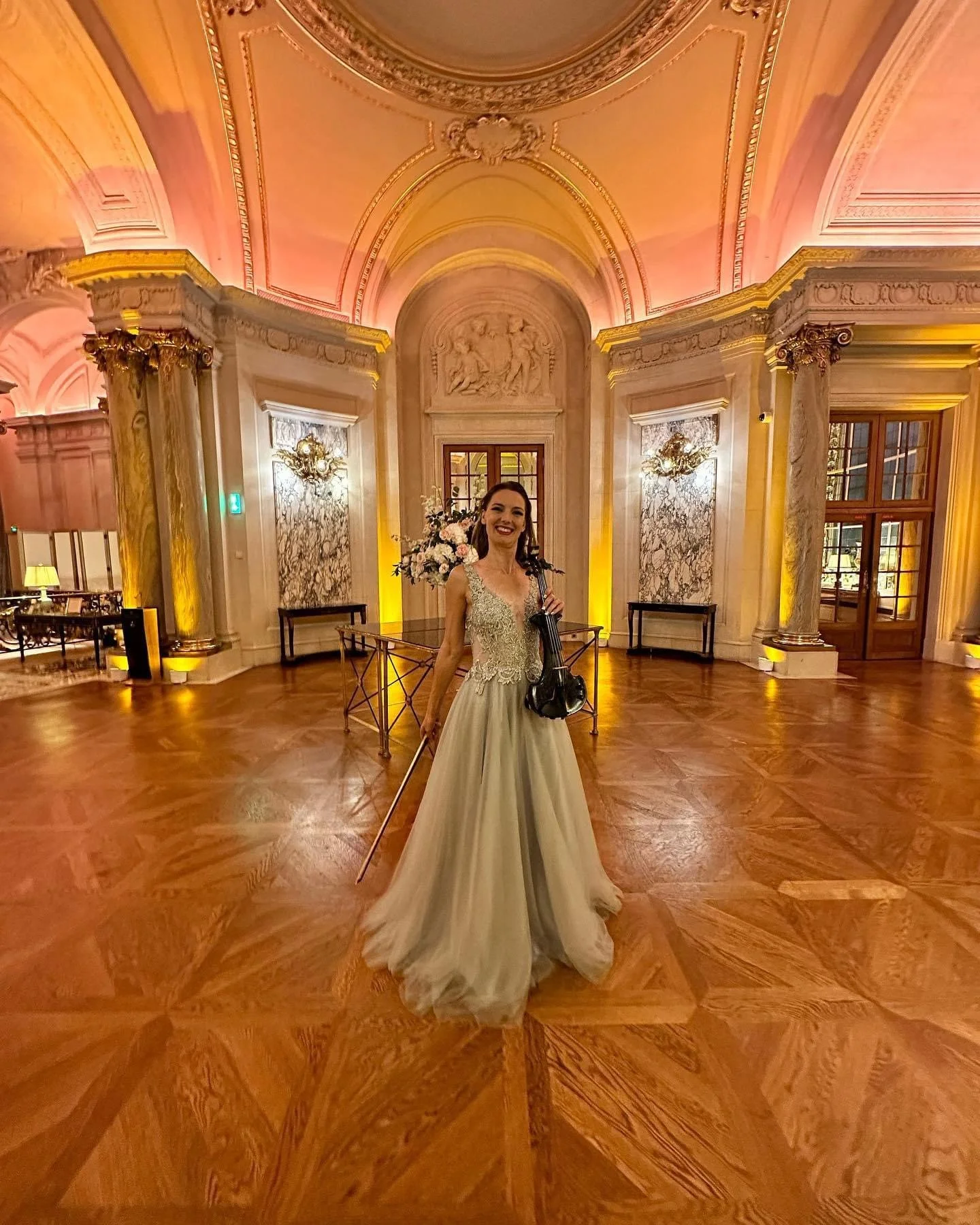 A woman in a wedding dress holding a violin and bow, smiling, standing in an ornate, elegant hall with marble columns, floral arrangements, and intricate ceiling details.