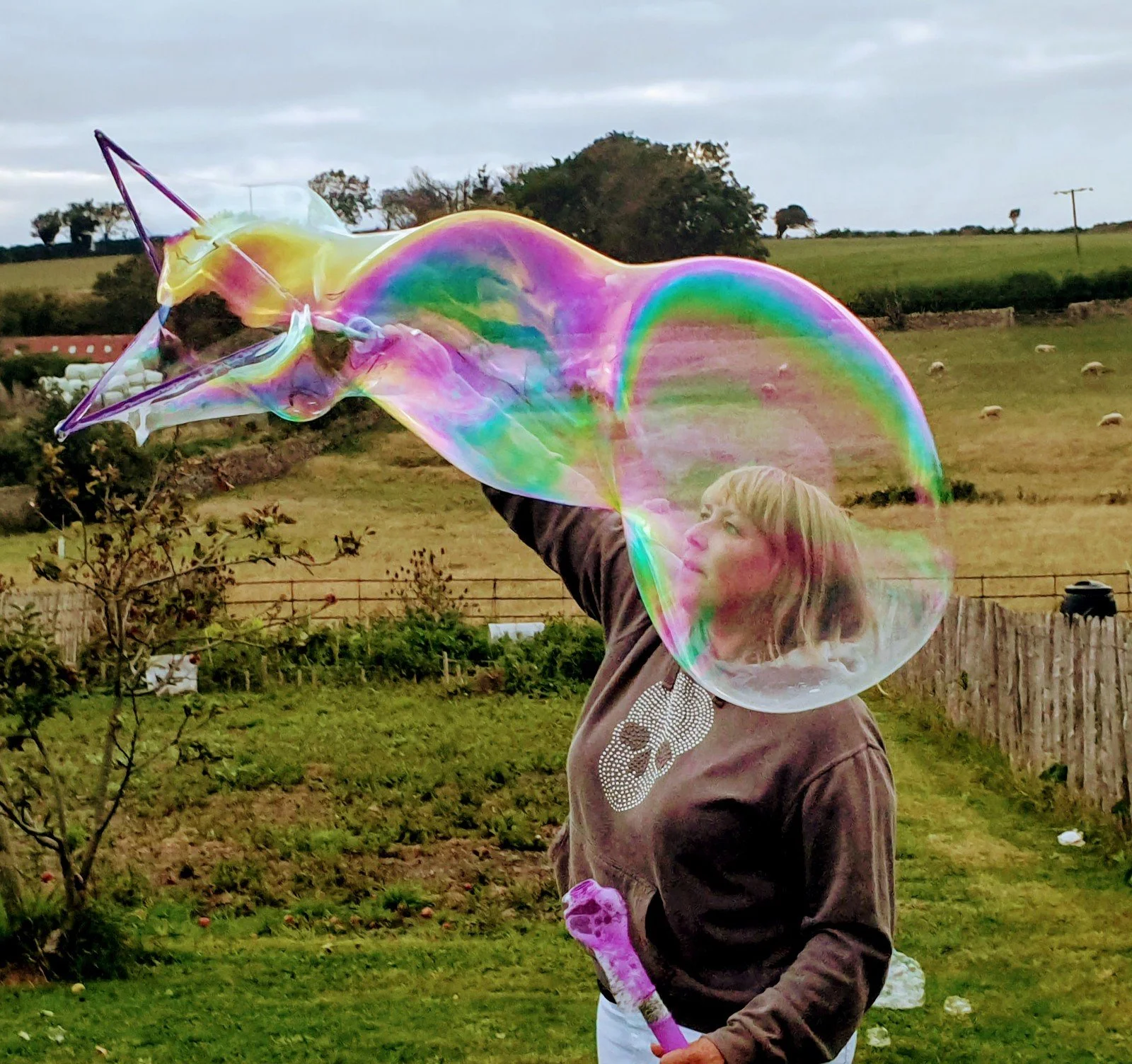 A person with blond hair and glasses holding a large, iridescent soap bubble outdoors in a grassy area with trees and a fence.