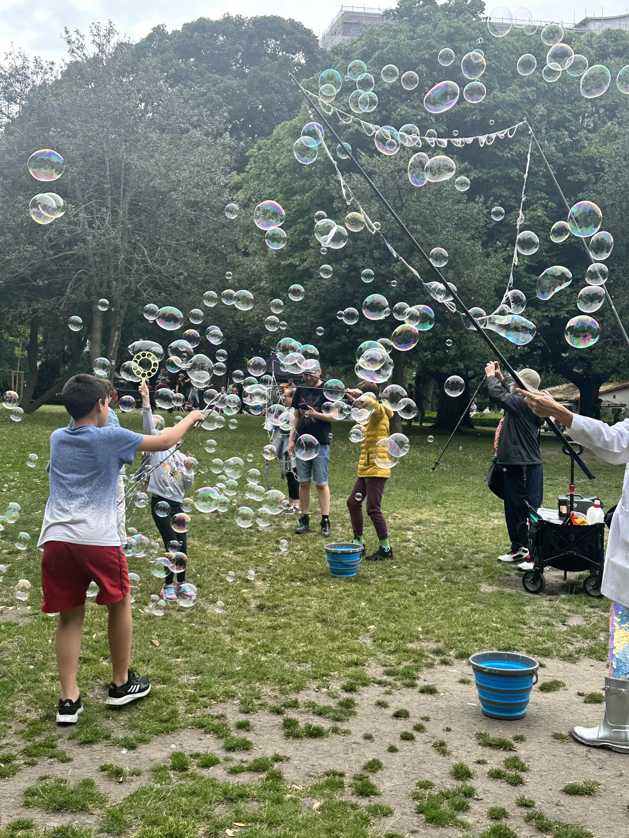 Children and adults in a park playing with soap bubbles, some holding bubble wands, with trees and a cloudy sky in the background.