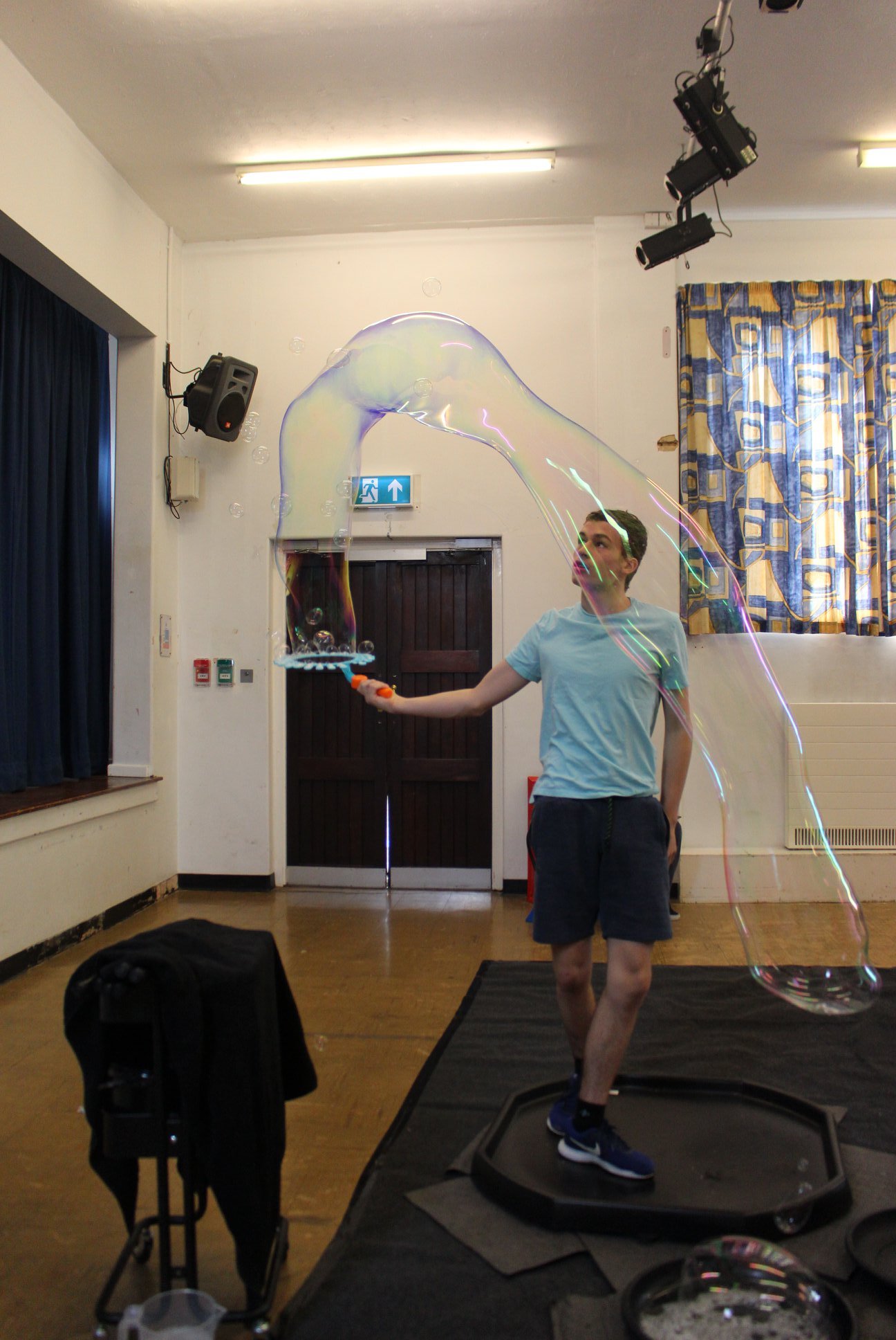 A young man is standing on a small trampoline, holding a bubble wand, creating large soap bubbles indoors. The room has wooden floors, a white wall with a blue and yellow curtain, and various equipment including a speaker, stage, and lighting.