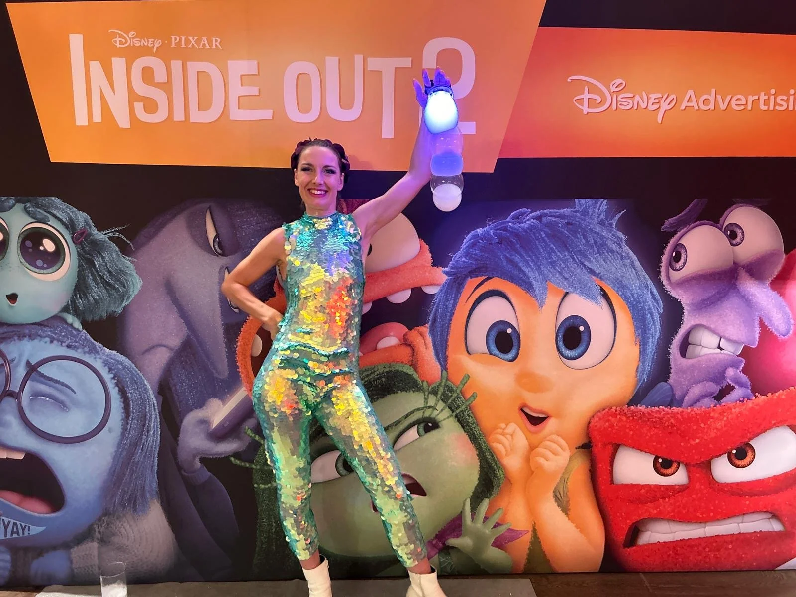 A woman in a shiny, multicolored, sequined jumpsuit stands in front of a promotional backdrop for the movie 'Inside Out', featuring several of the movie's character mascots. She is holding a bubble sculpture.