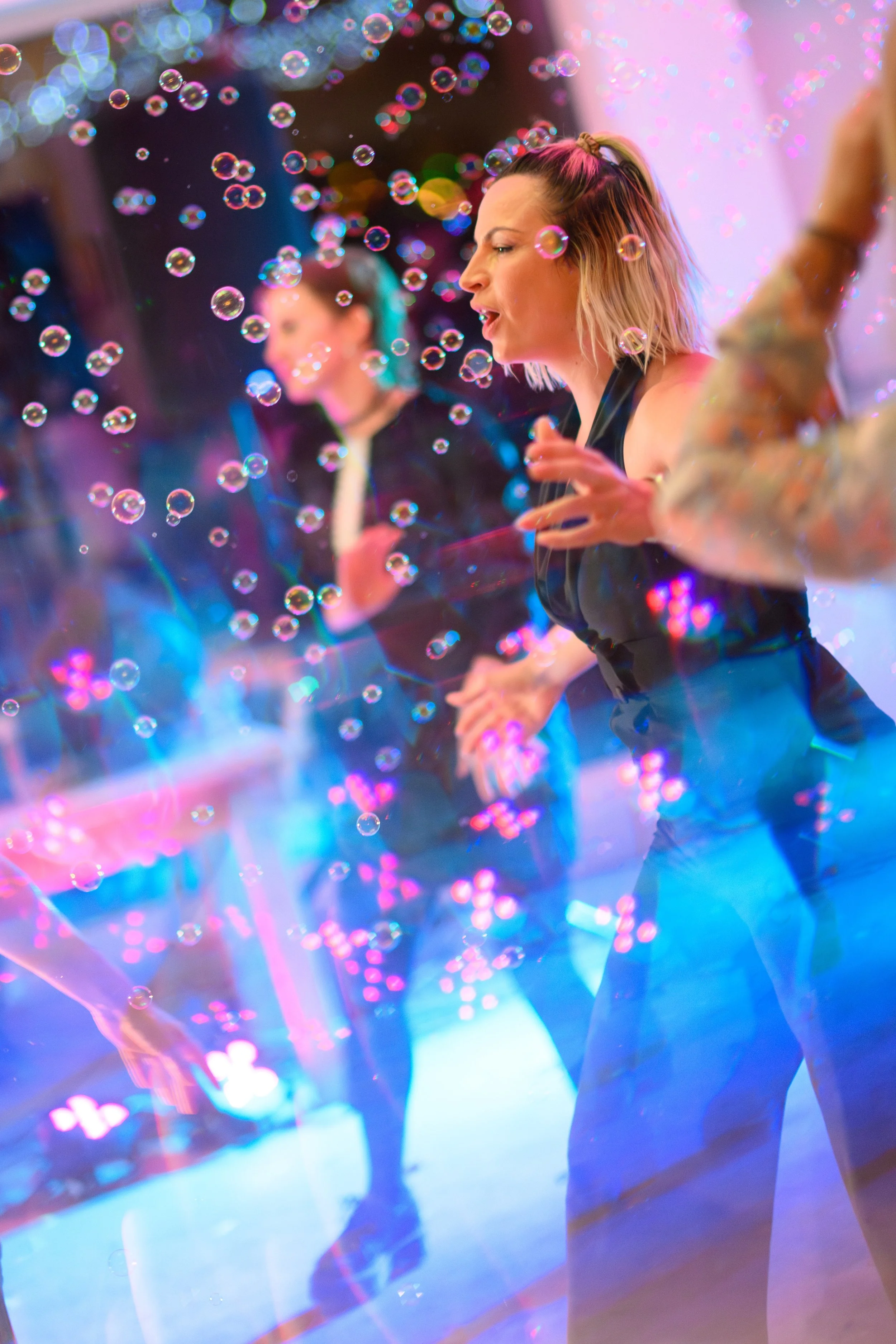 A woman with blonde hair in a ponytail is surrounded by colorful bubbles and neon lights, dancing on a vibrant, illuminated dance floor.