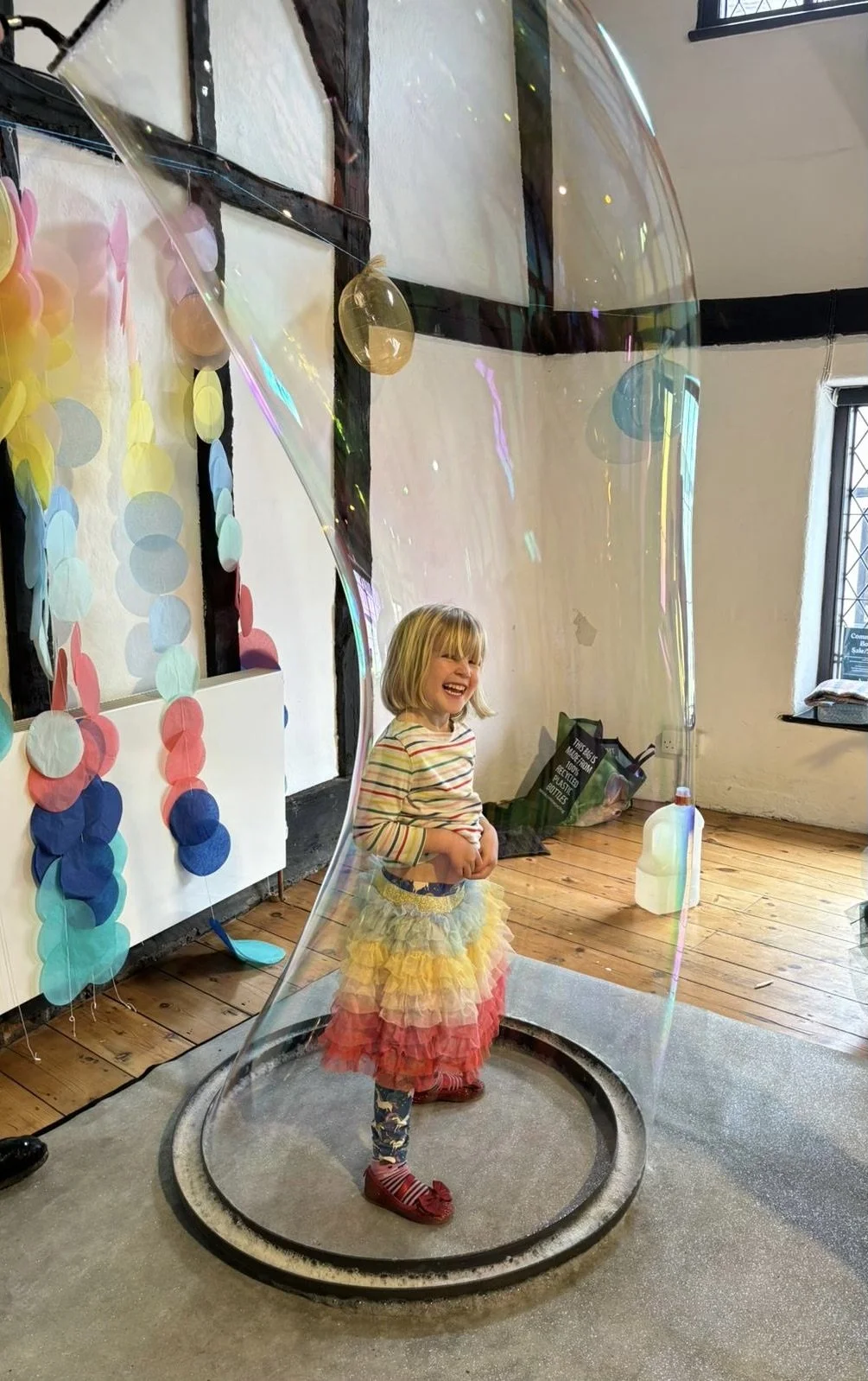 A young girl with a rainbow-colored ruffled skirt and striped top standing inside a large soap bubble, laughing indoors with colorful paper decorations on the wall.