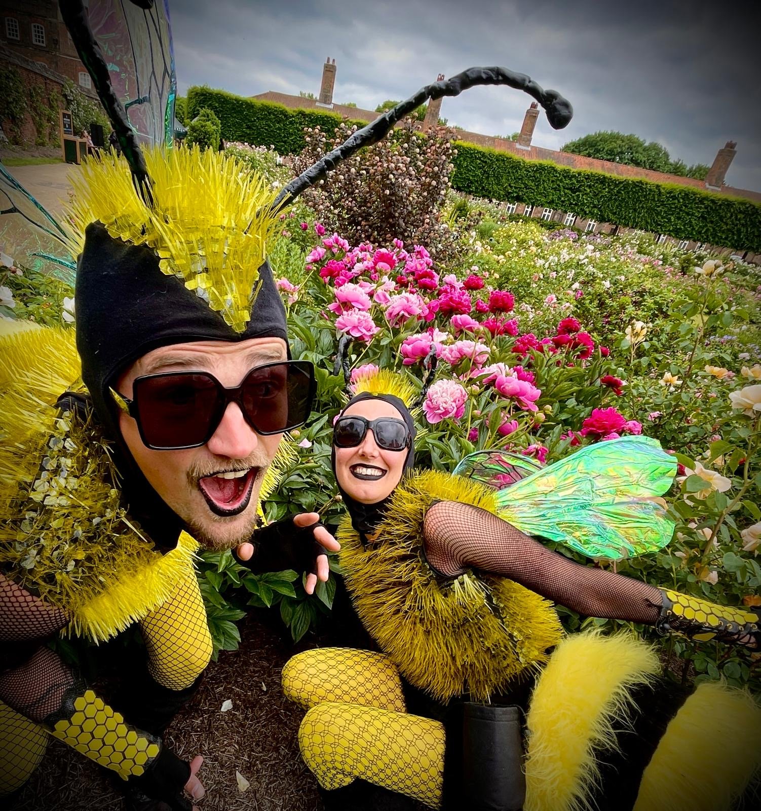 Two people dressed in bee costumes with yellow and black clothing, yellow wings, and antennae, taking a selfie in a garden filled with pink and white flowers under a cloudy sky.