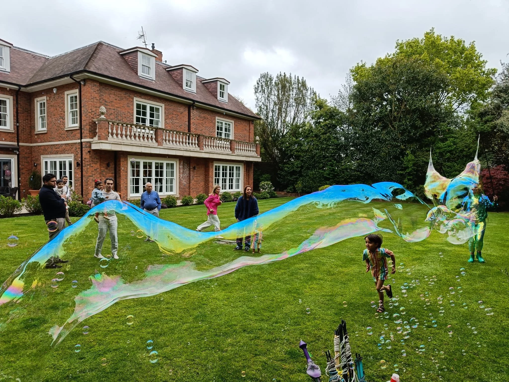 People playing with soap bubbles on a lawn in front of a large brick house with trees and a cloudy sky.