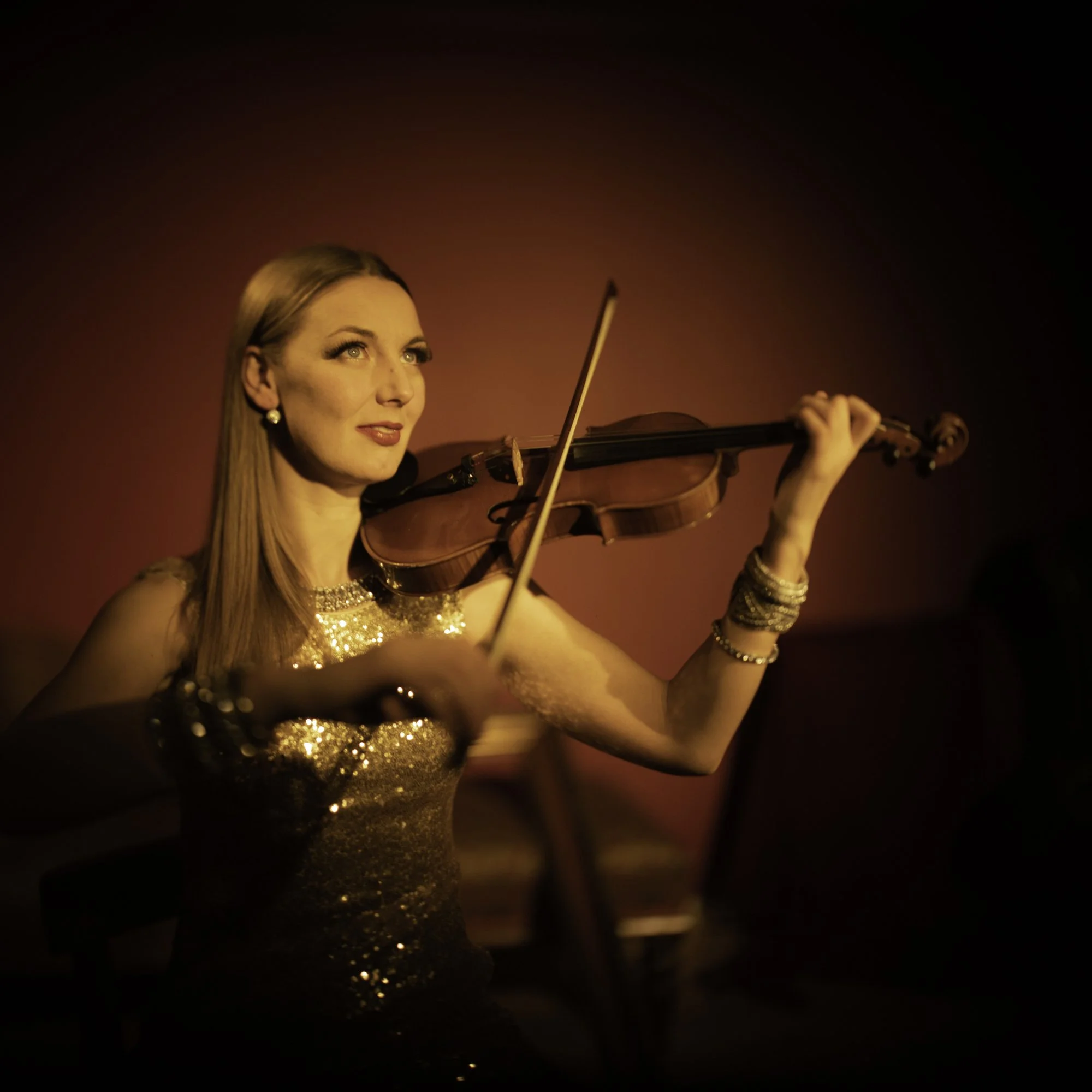 A woman with red hair playing the violin against a red wall background, wearing a sparkling gold dress and jewelry.