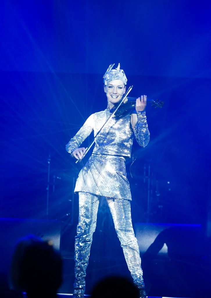 Performer dressed in a shiny, mirror silver costume with a matching crown, playing a violin on stage with blue lighting.