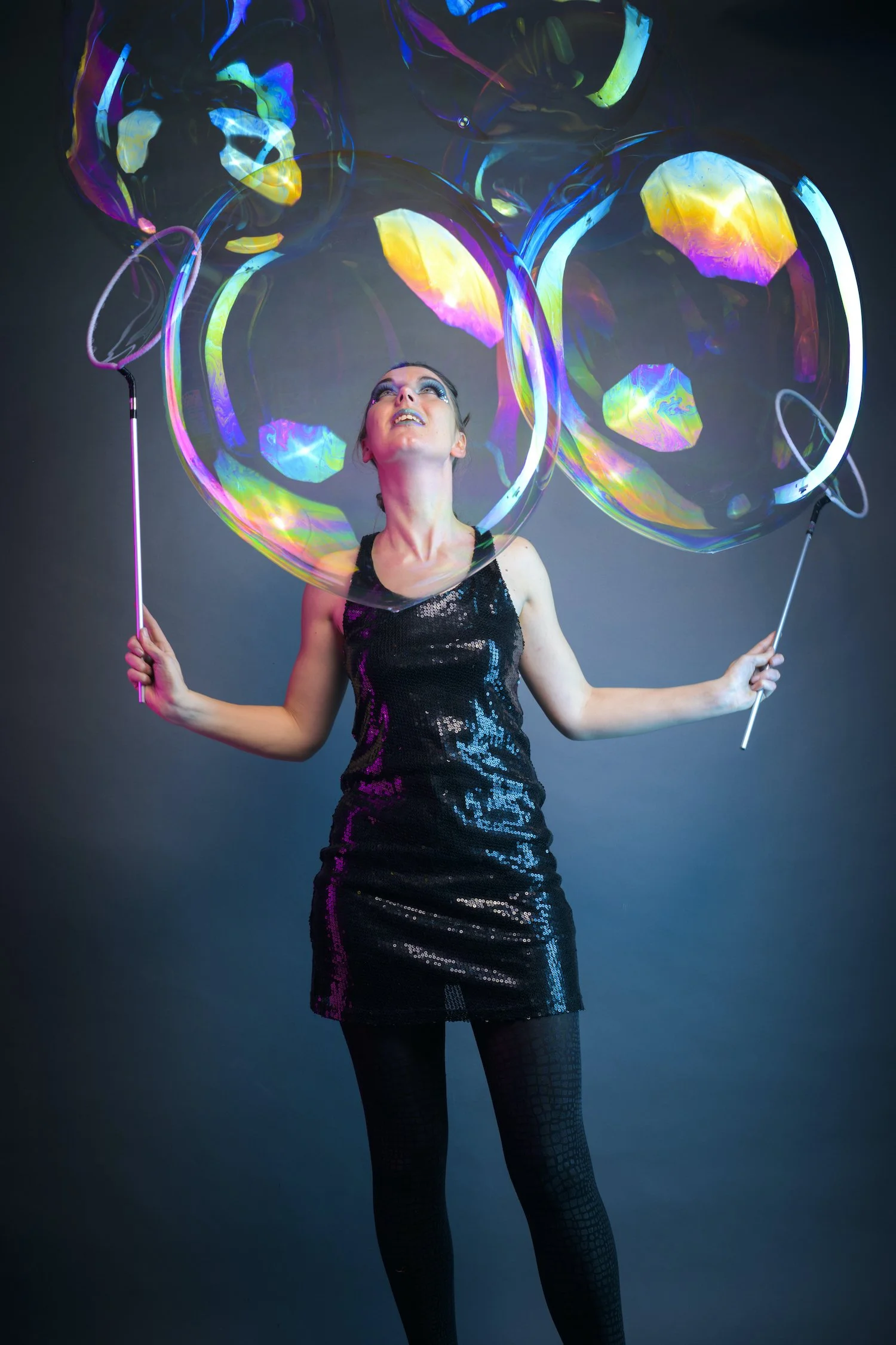 A woman in a black, shiny dress and fishnet stockings holds two large bubble wands with iridescent bubbles floating around her against a dark background, looking upwards.