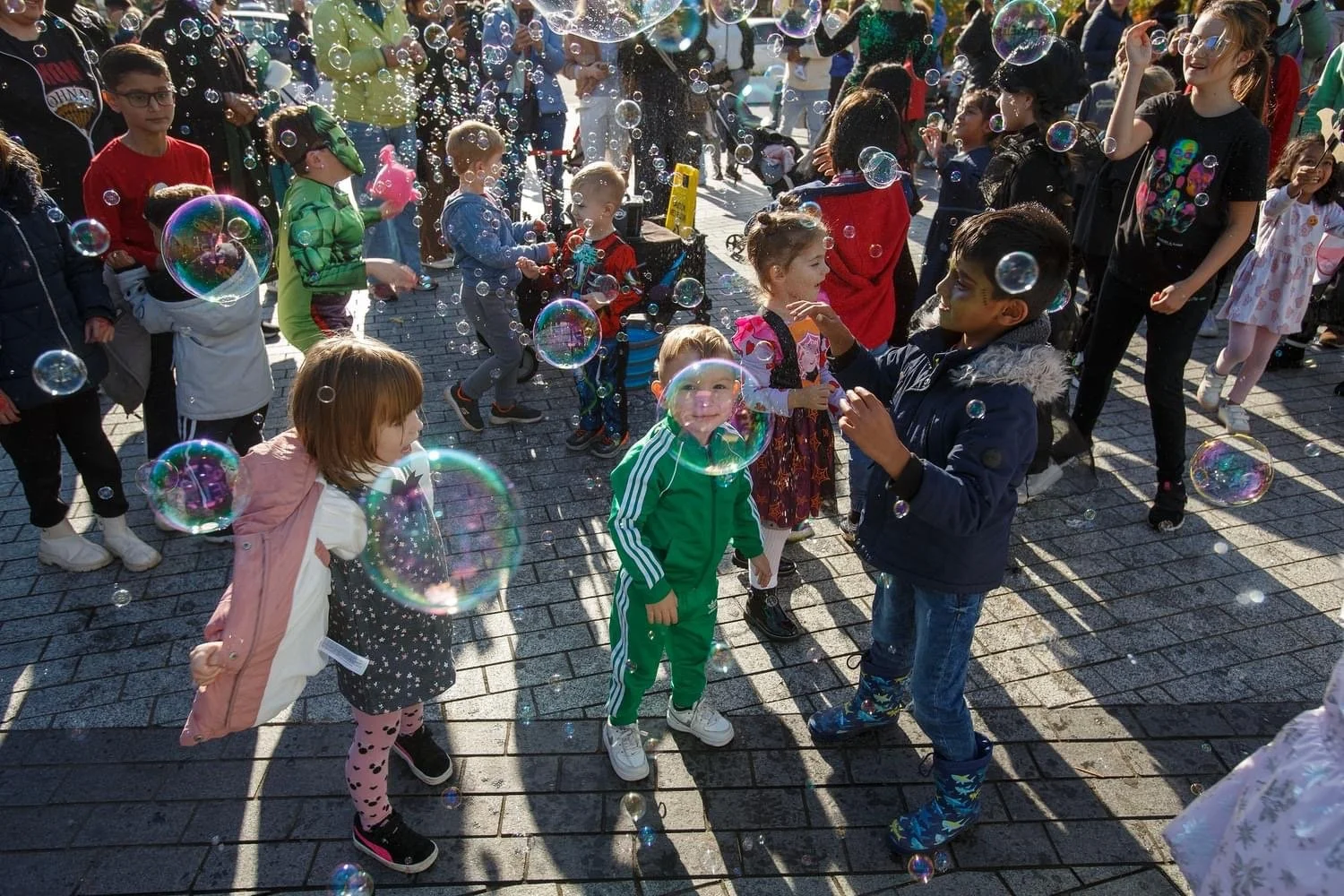 Children playing outdoors among bubbles at a lively event with many onlookers.