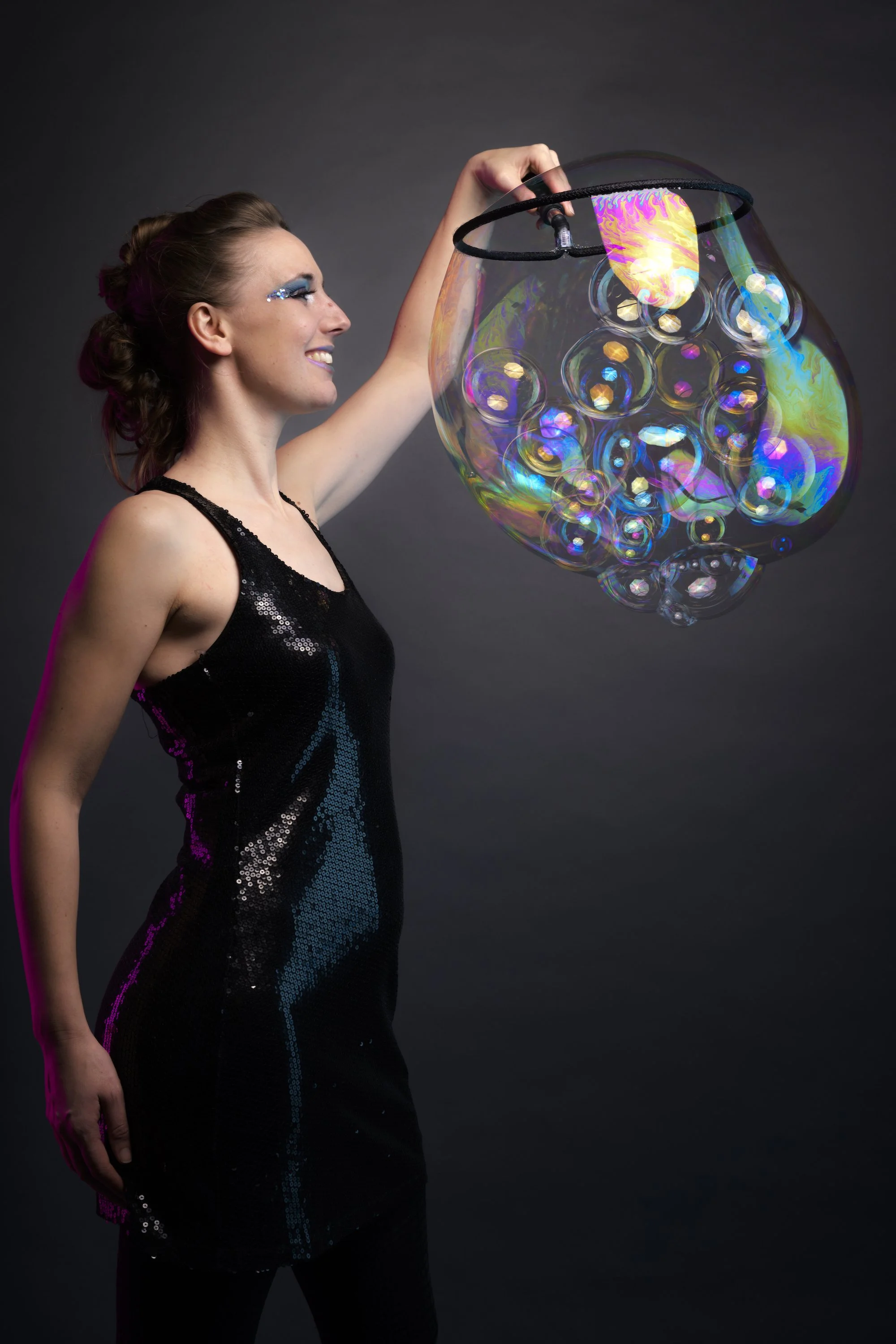 A woman with curly hair and blue eye makeup, wearing a black dress with sequins, smiling and holding a large iridescent soap bubble