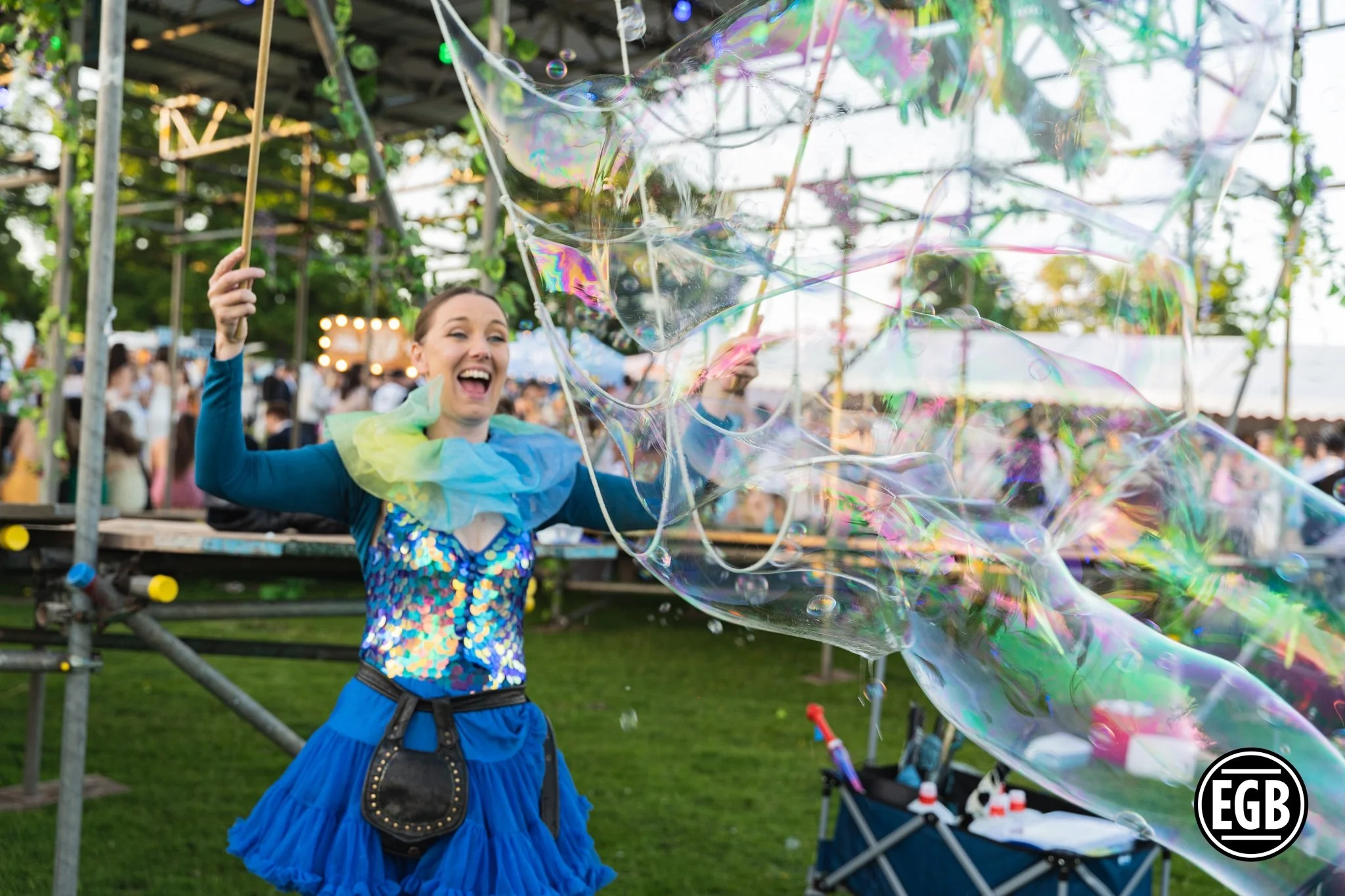 A woman in a blue skirt and colorful top is smiling and playing with large soap bubbles at an outdoor event or festival, with a crowd in the background and greenery around.