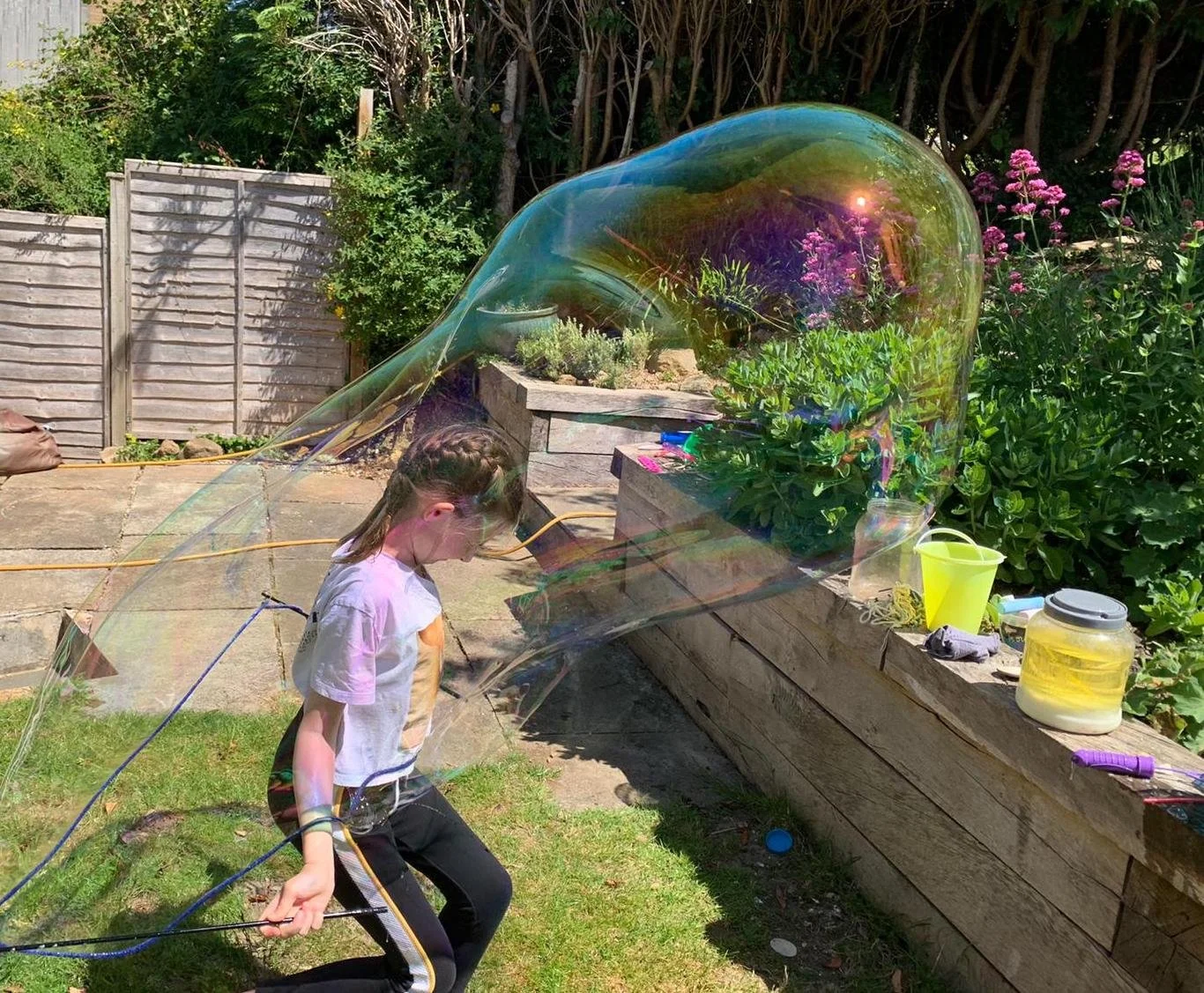 A young girl is walking in a garden with large soap bubbles floating above her. She is wearing a white T-shirt, black pants with a yellow stripe, and has braided hair. The background features a wooden fence, green bushes, and pink flowers, with gardening tools and supplies on a wooden ledge.