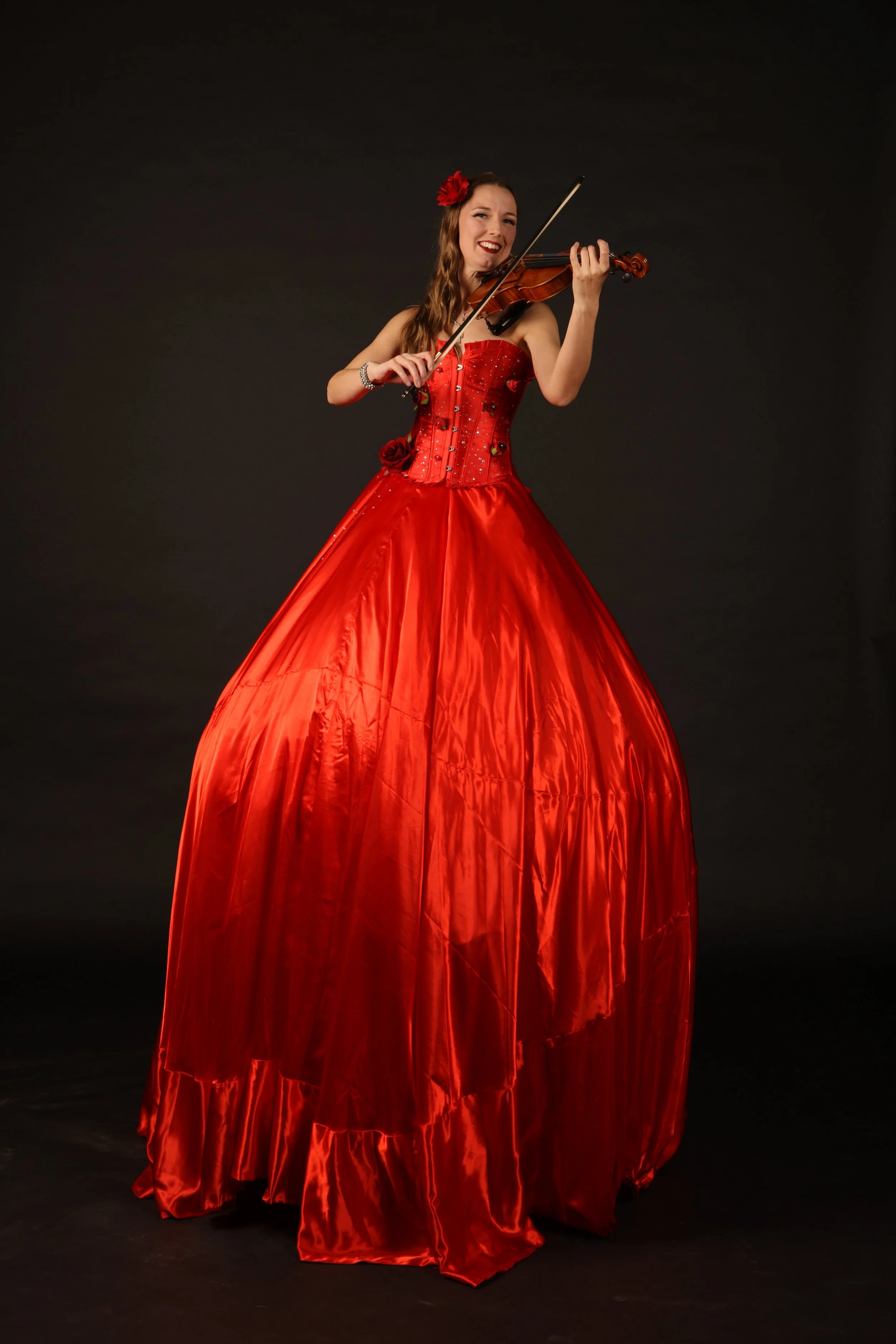 A woman on stilts in a vibrant red gown playing a violin against a black background.
