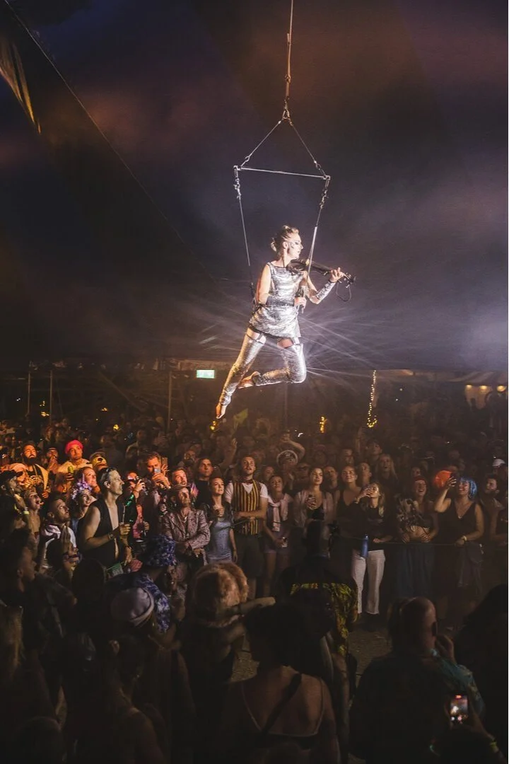A female performer in a silver outfit playing the violin while suspended in midair on a trapeze during a night show, surrounded by an audience of diverse spectators.