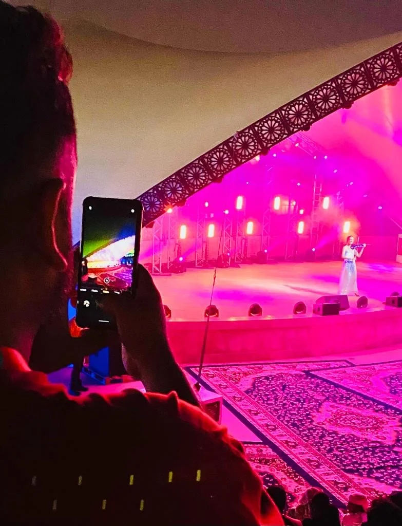Person in red shirt taking photo with a smartphone of a performer on stage at a colorful, pink-lit concert or show with intricate carpeted floor and stage lighting.
