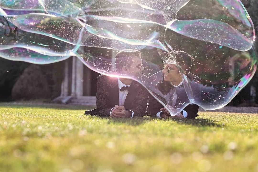 Two men in tuxedos lying on grass, looking at each other, with large soap bubbles floating overhead.