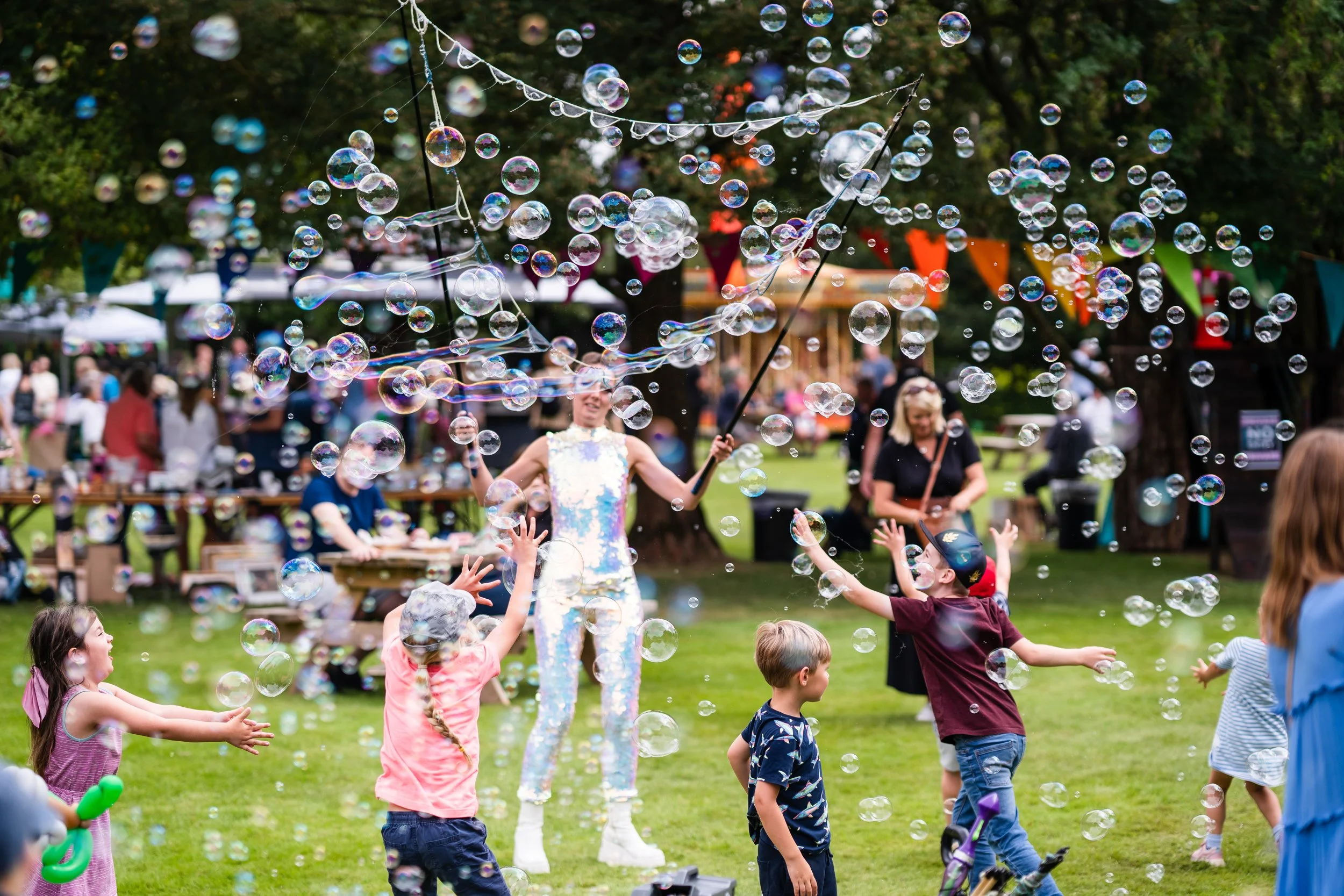 Children play with bubbles at an outdoor festival or park, with a woman in a colorful outfit creating the bubbles. People and colorful banners are visible in the background.