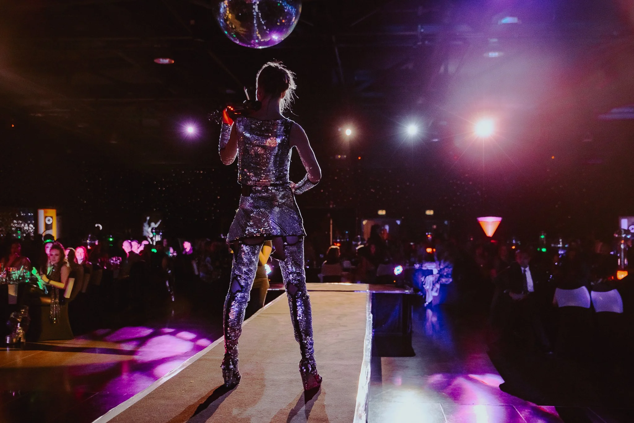 A performer in a shiny, metallic outfit on stage with audience and colorful lights in a nightclub or concert venue.